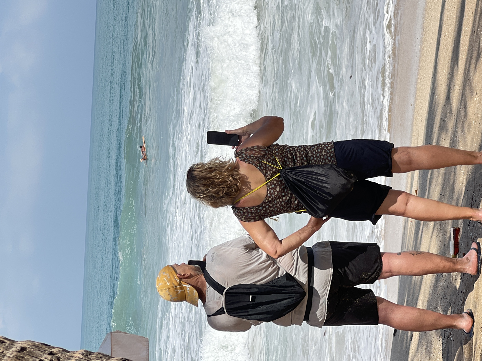 Two individuals, a male and a female, stand on a sandy beach facing the ocean in Kuta, Indonesia. The male, wearing a yellow baseball cap and a light-colored t-shirt over black shorts, has a black backpack slung over his shoulders and holds a phone in his right hand, seemingly taking a photo or video. The female, with curly blonde hair, wears a patterned top and dark shorts, also holding a black smartphone in her right hand, her gaze directed towards the water. In the distance, two individuals are seen in the ocean, one on a surfboard and another in the water. The waves are breaking, creating white foam that reaches the shore. The sky is partly cloudy, and shadows from palm trees or other foliage are cast on the sand.