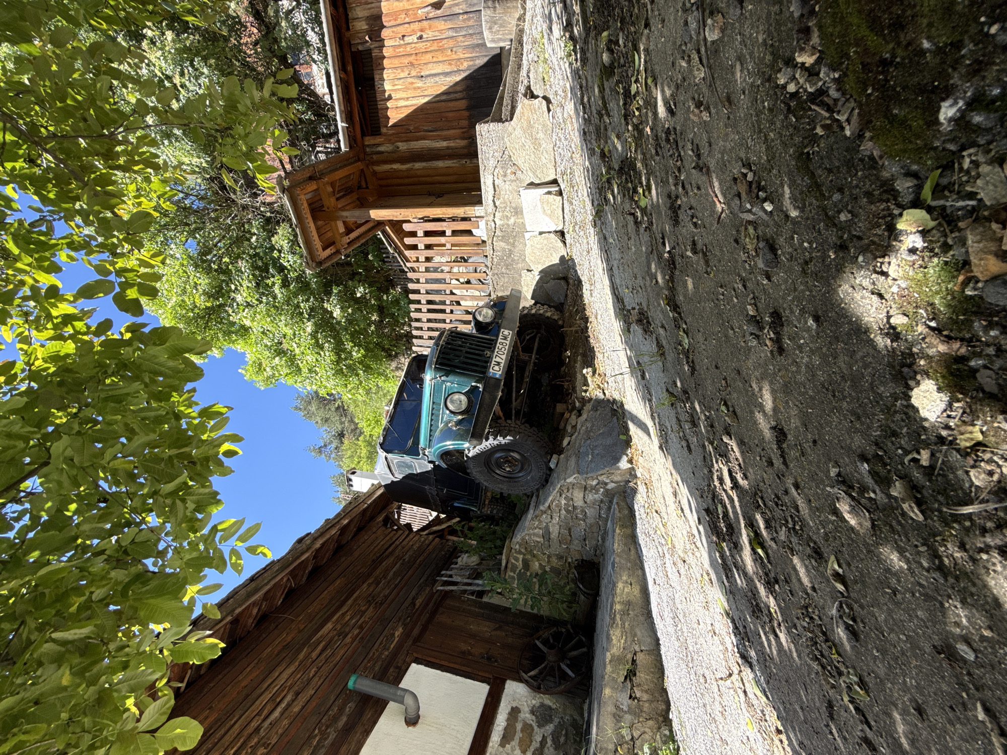In Shiroka Laka, Bulgaria, an olive-green vintage off-road vehicle, identified by its license plate "CM 7058 MC", is parked on a steep, gravelly incline adjacent to a rustic wooden building. The vehicle is positioned on a stone foundation, with its front tires angled towards the left and its rear tires resting on the ground. The building, constructed of weathered wooden planks, features a prominent pitched roof and an attached wooden shelter. Nearby, a wooden fence encloses an area to the right of the building. A traditional wooden wheel, possibly an antique artifact, lies on the ground to the left of the building's entrance. The scene is bathed in sunlight, casting shadows from the surrounding trees and foliage onto the ground and structures. The sky is clear and blue, with the lush green canopy of trees overhead dappling the scene with light and shade. The ground is a mixture of gravel, dirt, and scattered stones, indicating a rural or semi-rural setting.