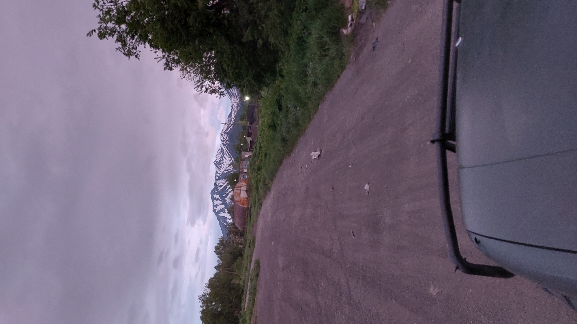 The image depicts a dirt road leading towards a cluster of buildings at the base of snow-capped mountains under a cloudy, dusky sky. The perspective suggests the photo was taken from a vehicle, as the hood and a portion of the roll cage are visible in the foreground. The environment appears rural, with greenery bordering the road and scattered debris on the road surface. The buildings in the middle ground seem somewhat dilapidated, with one structure appearing to be an abandoned greenhouse. The overall lighting indicates either dawn or dusk. The location is identified as the city of Лесной, Russia.