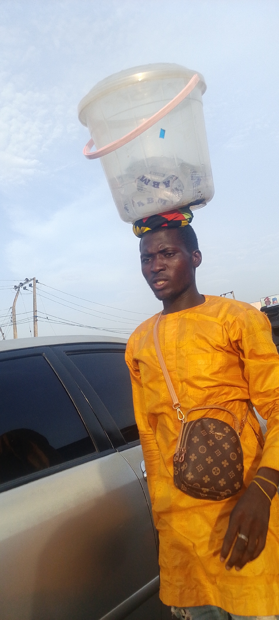 A young man is depicted outdoors, likely in Lokoja, Nigeria, under a bright, cloudy sky. He is carrying a transparent plastic bucket with a pink handle on his head, balanced on a colorful patterned cloth. The bucket contains multiple clear plastic sachets, primarily bearing the visible text "ABM" and "Water," suggesting he is selling sachet water or a similar beverage.
The man is of African descent, wearing a bright yellow, long-sleeved traditional tunic. He has a small, patterned crossbody bag slung over his shoulder, which features a design similar to luxury brand monograms. Yellow string bracelets are visible on his right wrist, and a ring is on his left ring finger. His expression is neutral or slightly pensive, and he looks towards the viewer's left.
Part of a silver or light-colored car is visible beside him, indicating a street or roadside setting. In the background, utility poles and a dense network of overhead electrical wires are present. The lighting suggests it is daytime, possibly morning or late afternoon. The scene captures a moment of daily life, likely involving street vending.