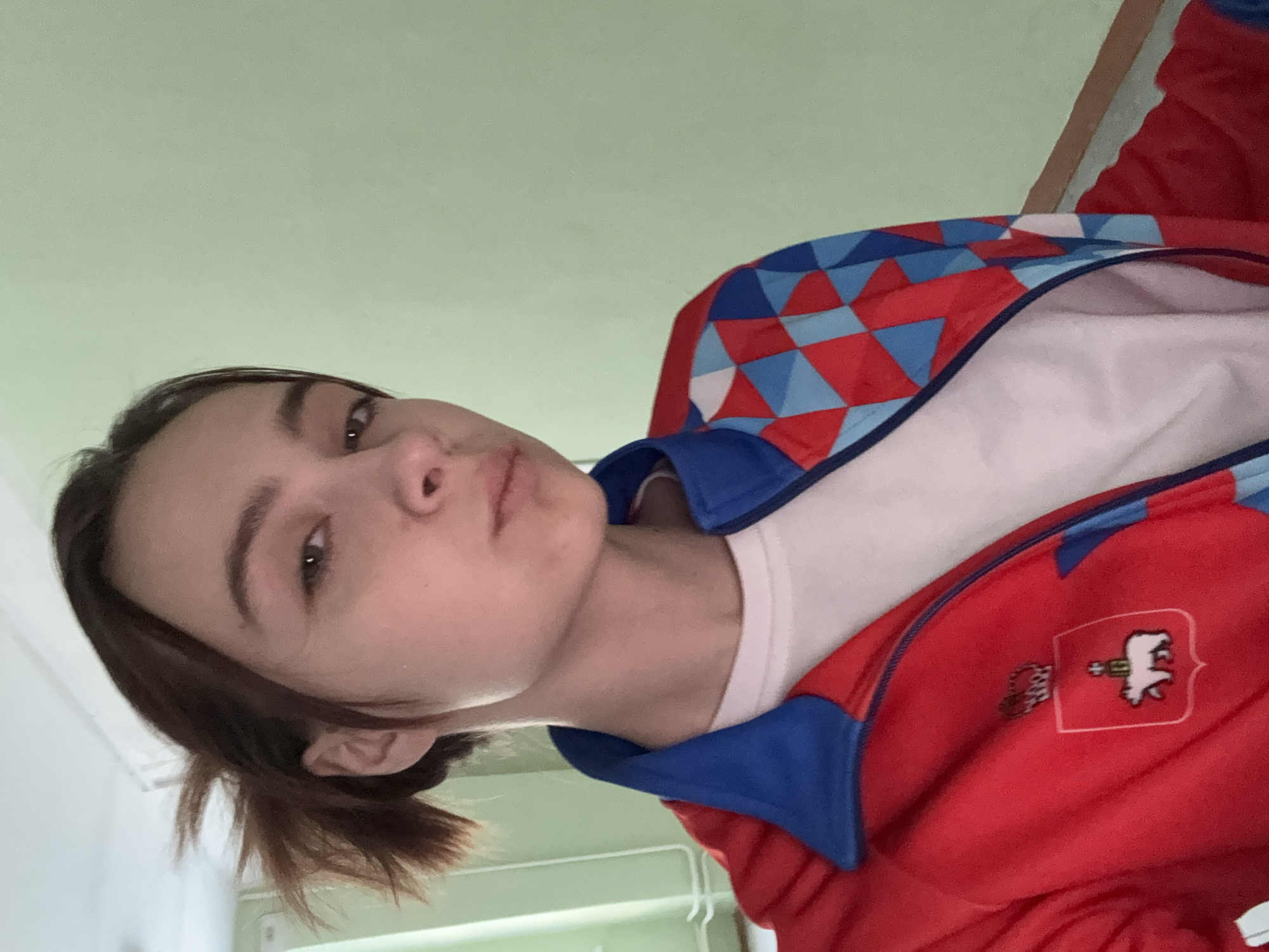 A close-up, slightly angled selfie of a young woman with short brown hair, wearing a white t-shirt under a red jacket with blue trim and a patch featuring a white bear emblem. The background is a plain, light green wall. The location may be Berezniki, Russia, based on the emblem on the jacket.