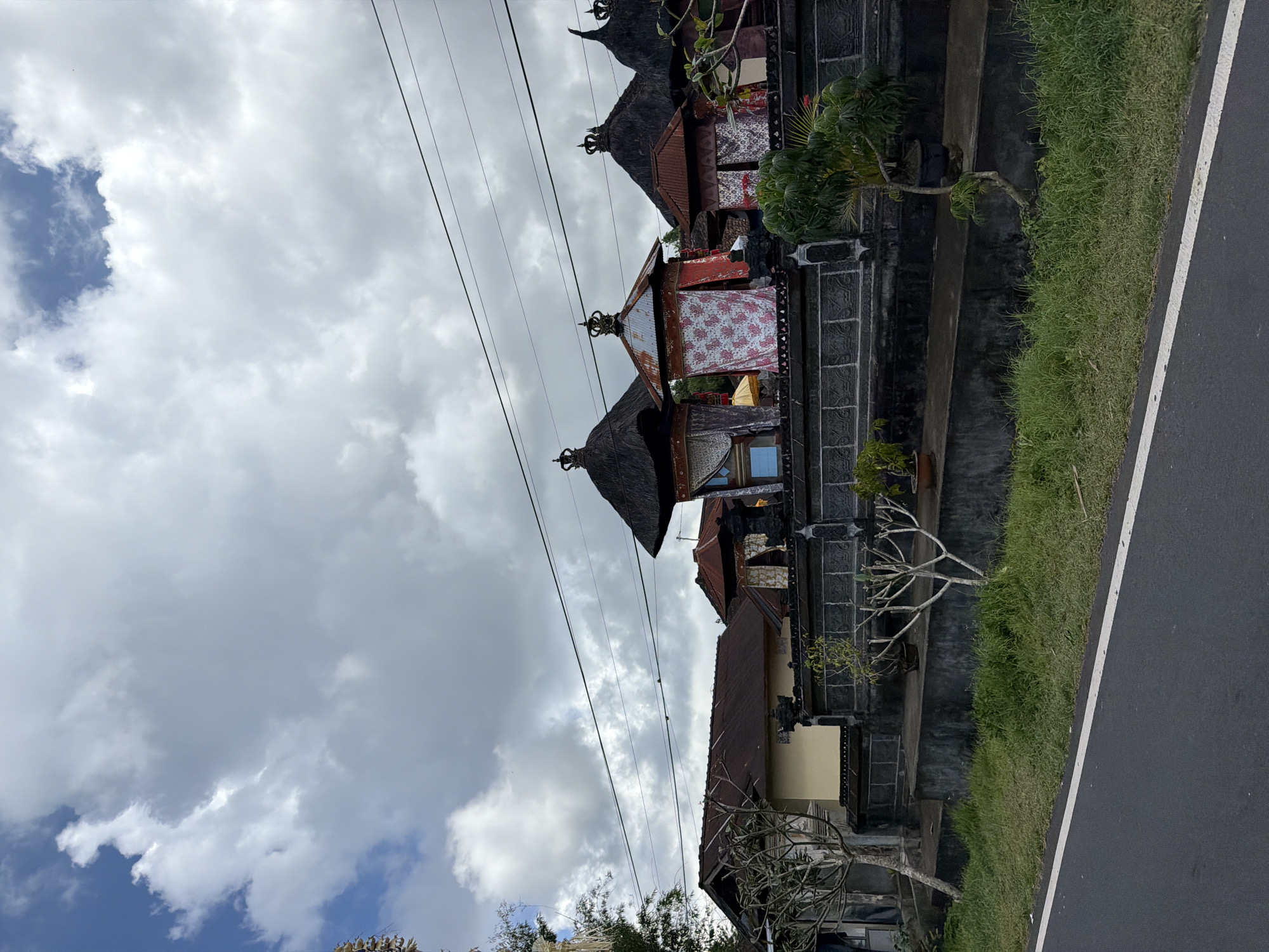 A three-quarters shot, taken from a roadside, shows a row of traditional Balinese buildings with thatched roofs and elaborate facades, adorned with colorful fabrics and crowns. The buildings are set behind a dark stone wall, with small potted trees and bushes lining the foreground. A paved road with a white line runs along the bottom of the frame, bordering a strip of green grass. The sky is filled with dramatic, puffy clouds. The scene is located in Susut, Indonesia.