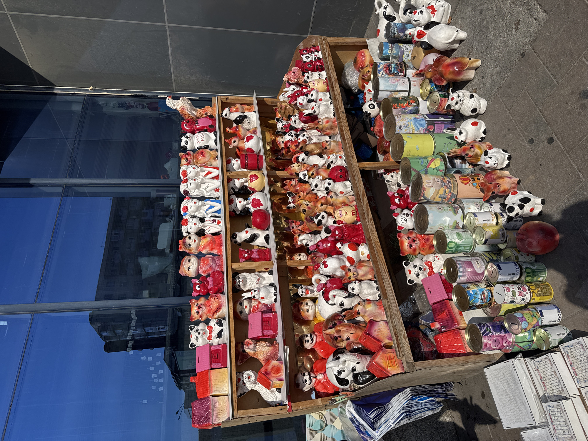 An outdoor stall displays an assortment of ceramic animal figures and piggy banks. The stall is set up in front of a large glass-windowed building, with a dark grey tiled wall to the right. The ground is paved with grey flagstones. In the background, a multi-story building is visible through the glass. The ceramic figures include various animals such as cats, dogs, rabbits, and pigs, many painted in bright colors. Several are shaped like houses and appear to be piggy banks. Stacked cylindrical coin banks are also present, some decorated with cartoon characters. The scene is bathed in sunlight, casting shadows on the ground, suggesting it is daytime. No people are visible in the immediate vicinity of the stall, but the overall context suggests a market or street vendor setting. The presence of coin banks in various denominations, some featuring euro and dollar symbols, may indicate their intended market. The specific location in Baku, Azerbaijan, is not directly evident from visual cues within the image itself, but the style of the goods and the architectural elements could be characteristic of the region.