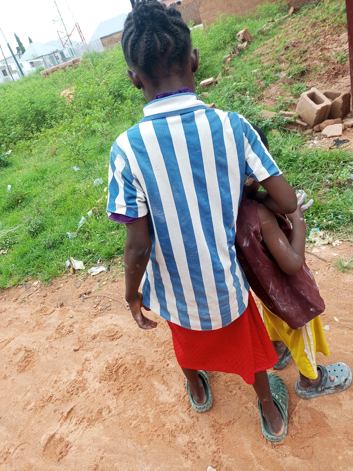 In Tempure, Nigeria, a person stands on sandy ground, viewed from behind. They wear a blue and white striped shirt and a red skirt. Another person is partially visible, embraced by the first. The environment includes overgrown green grass and some scattered debris, with buildings and a cell tower in the background. The lighting suggests it is daytime.