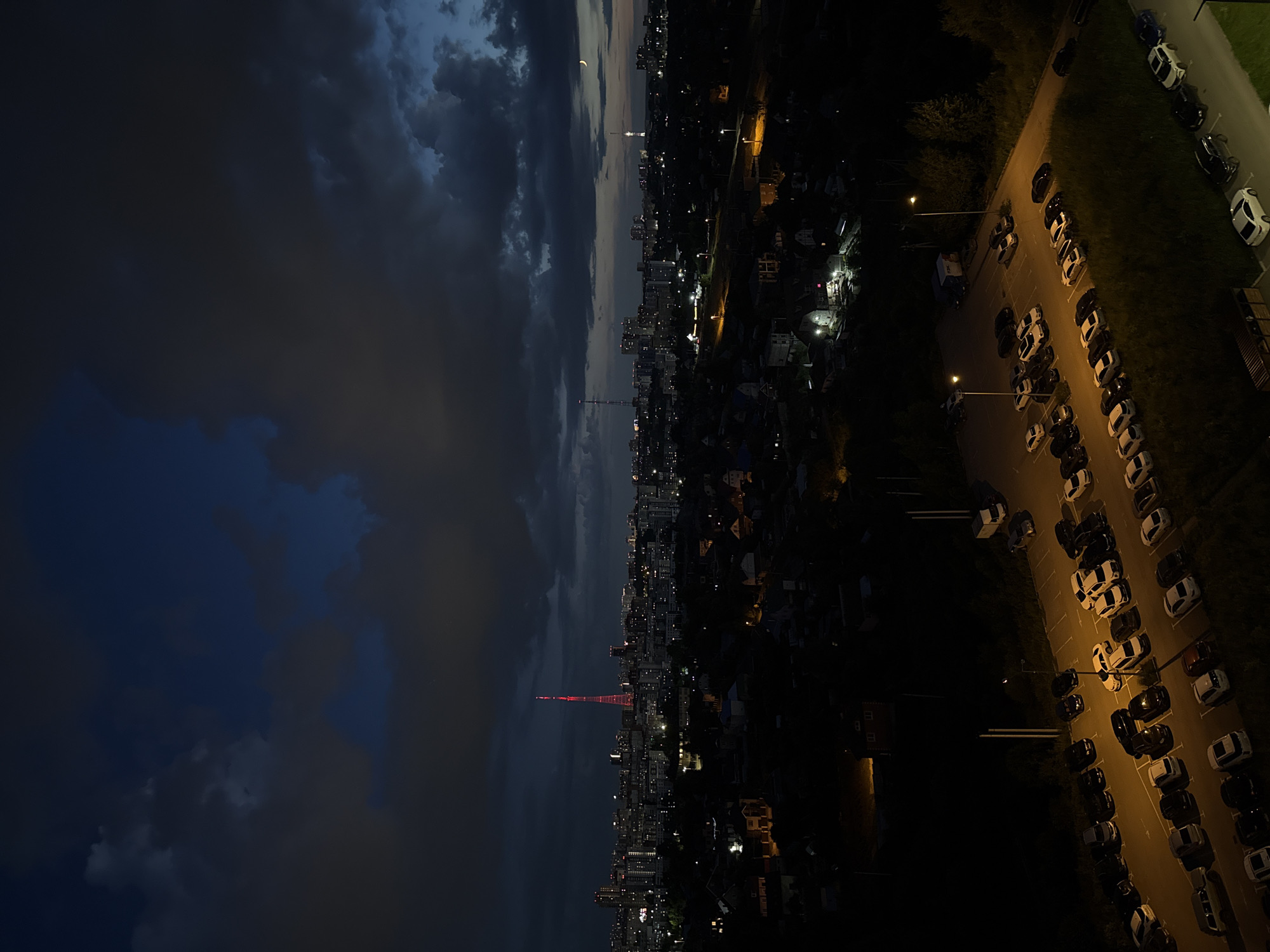 The image captures a nighttime cityscape, likely Perm, Russia, characterized by a dark sky with scattered clouds and the faint glow of city lights. A prominent red-lit tower, possibly a television tower, stands out against the urban sprawl. Residential areas with illuminated houses and streets are visible in the foreground, transitioning into a more densely built-up city center. A parking lot filled with numerous cars is illuminated by streetlights, creating a warm orange glow on the asphalt and vehicles. The overall scene conveys a sense of a quiet evening in a moderately sized city.