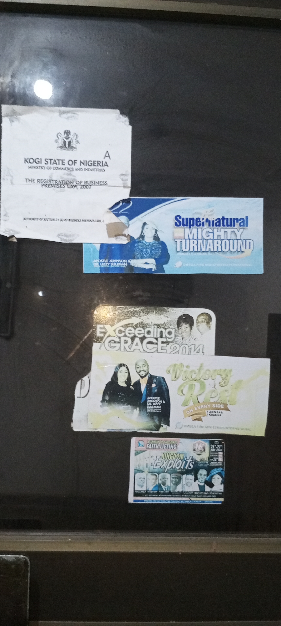 The image depicts a collection of posters and documents affixed to a dark, possibly metallic, surface. At the top is a document titled "KOGI STATE OF NIGERIA MINISTRY OF COMMERCE AND INDUSTRIES THE REGISTRATION OF BUSINESS PREMISES LAW, 2007." Beneath this is a poster for "Supernatural MIGHTY TURNAROUND" featuring Apostle Johnson & Dr. Lizzy Suleman of Omega Fire Ministries International. Below, is a partially obscured poster advertising "Exceeding GRACE 2014", again featuring Apostle Johnson & Dr. Lizzy Suleman. Next, there is a poster for "Victory REST ON EVERY SIDE" of Omega Fire Ministries International, citing scripture from 1 John 5:4 & 1 Kings 5:4. At the bottom is a poster for "FAITH LIFTING KINGDOM Exploits." The posters appear to be promotional materials for religious events. The location is implied to be Adankolo, Nigeria.