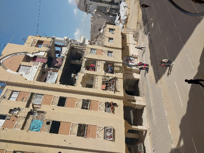 The image depicts an urban street scene in Khan Yunis, Palestinian Territory, under a clear blue sky with sparse clouds. A multi-story, light-colored building, exhibiting extensive damage, occupies the left and center of the frame. The building features numerous broken windows and visible impact marks across its facade. Significant structural damage is concentrated on the right side, where large sections of walls and balconies are missing, exposing interior spaces. Despite the damage, laundry hangs from several balconies. On the ground floor, a silver sedan is parked beneath an arched recess.
A paved road runs horizontally across the mid-ground, flanked by a paved sidewalk. On the sidewalk near the damaged building, a group of approximately five individuals, including adults and children, are clustered. A smaller group of three children stands on the paved road further to the left. In the lower-left foreground, a solitary adult figure, clad in dark clothing, walks on the street away from the viewer. Another individual walks on the street in the mid-ground toward the right. The ground adjacent to the street and around the damaged structures appears dusty and contains scattered debris piles. In the background, further down the street to the right, a dome-topped building with visible damage is present.