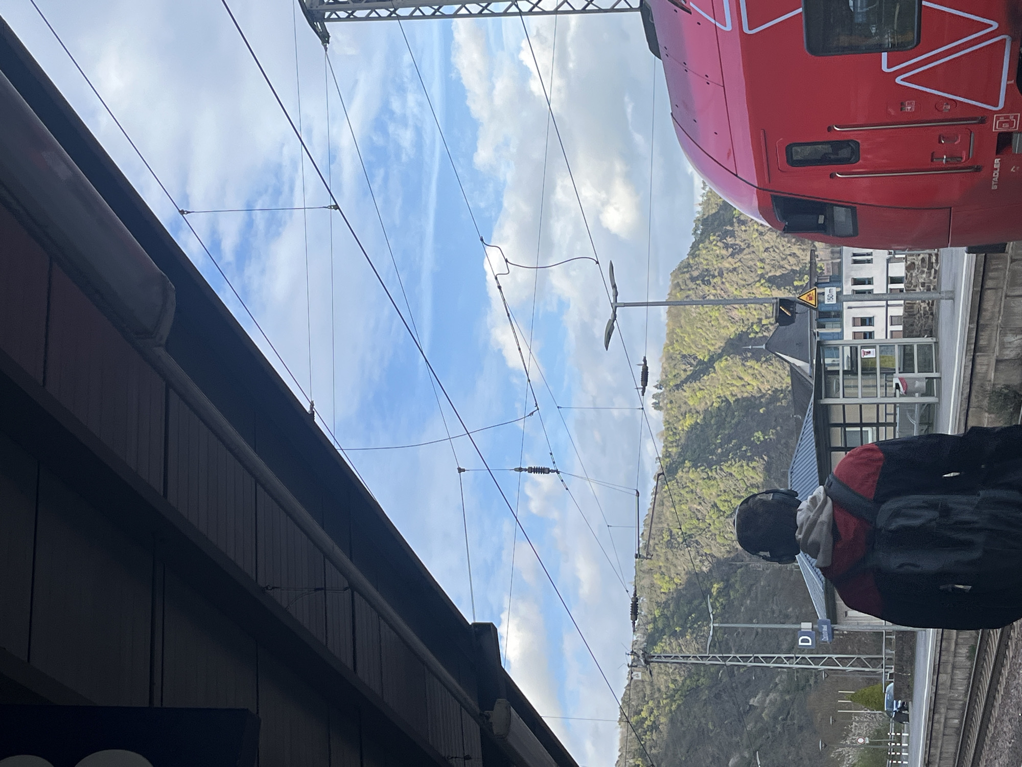 The image captures a scene at a train station in Cochem, Germany. The viewpoint is from a low angle, looking upwards towards a partly cloudy sky. Overhead electrical wires crisscross the view, indicating an electrified railway system. The edge of a modern red train is visible on the right, parked at the platform.
A person wearing a backpack and a jacket with red accents stands on the platform, facing away from the camera.
In the background, a hill covered with trees is visible. A building with multiple windows is located near the train. A blue sign with the letter "D" on it is also seen on the train platform. The photograph is slightly rotated.