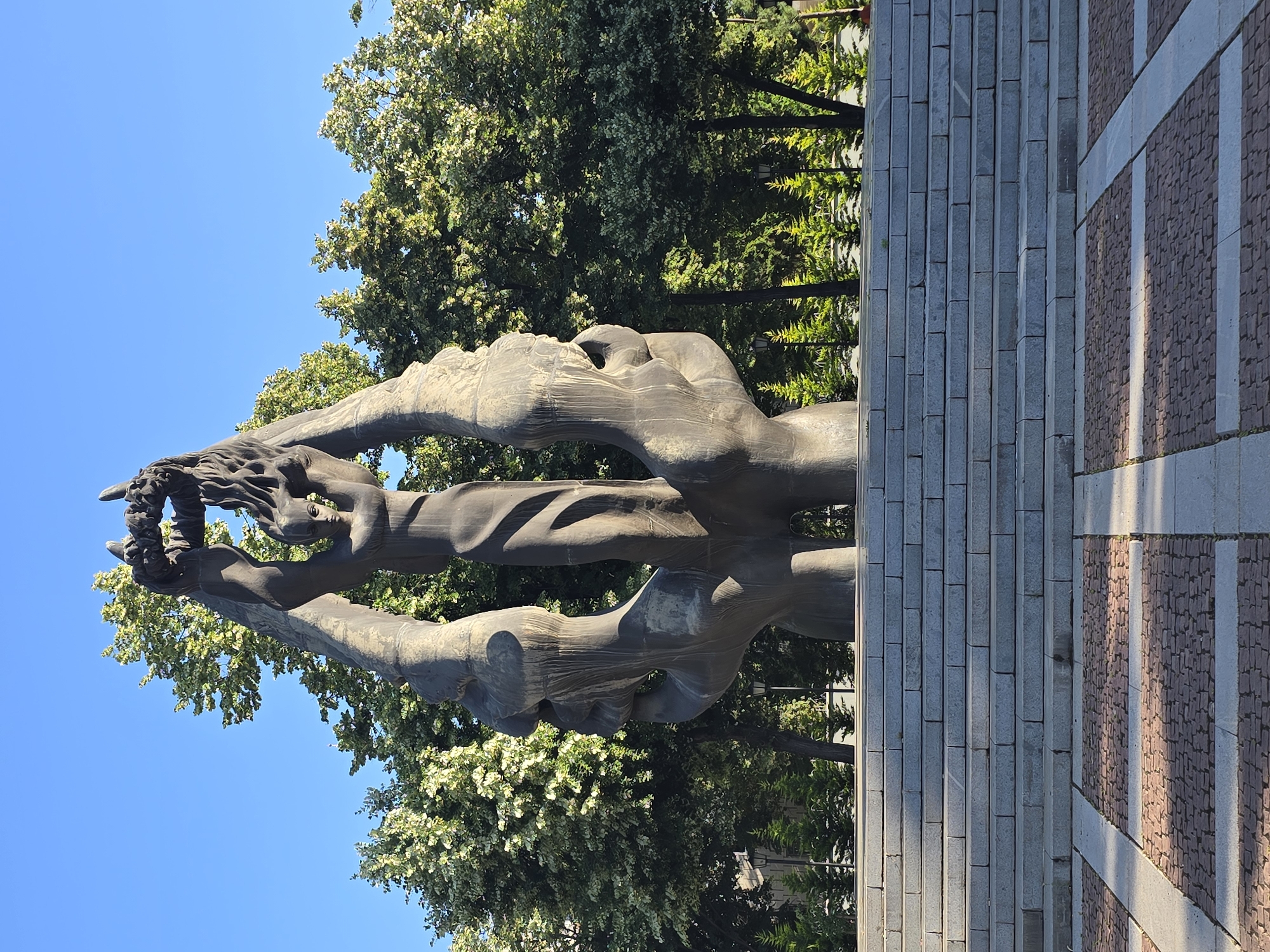 A monumental bronze sculpture depicting two large, stylized hands forming an arch over a figure of a woman holding a wreath. The sculpture is situated in front of a row of lush green trees, with a clear blue sky above. In the foreground, there are wide stone steps leading up to a plaza paved with red brick and stone. The overall scene suggests a public monument or memorial in an outdoor setting.