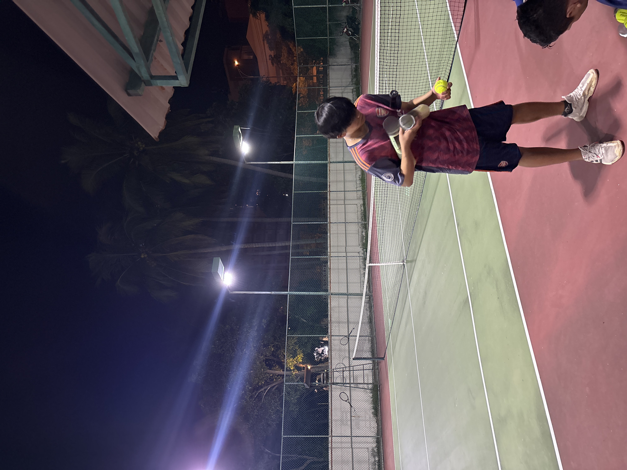 A young person stands on a tennis court at night, holding a tennis ball and a can of tennis balls. They are wearing a maroon and purple t-shirt with a team logo, dark blue shorts with a team logo, and light-colored sneakers. The tennis court is lit by overhead lights, illuminating the green and red surfaces and the net. Palm trees are visible in the background, along with a fence and buildings. Another person's head is visible in the lower right corner of the frame. The scene appears to be an outdoor tennis facility in an urban or suburban setting.