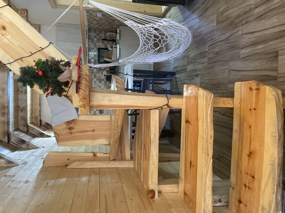 This image depicts an interior scene in Palež, Montenegro, characterized by a rustic and natural wood aesthetic. A prominent, light-toned, natural wood staircase with live-edge steps occupies the foreground and midground, curving upwards from the lower right to the upper left. Black string lights are wrapped around the central support post and banister of the staircase. A small artificial Christmas tree, adorned with red baubles and pinecones, rests on a flat wooden surface extending from the central staircase post, accompanied by a red felt decorative item. A light gray towel is draped over an upper section of the wooden staircase structure. The left wall is clad in horizontal light wood paneling. The floor is composed of variegated brown and gray wood-look tiles. In the background, a kitchen area is visible, featuring a light-colored square-tiled backsplash, white lower cabinets, a sink with a black tray holding items, and a visible cooktop. A white macrame-style hammock chair is suspended from a horizontal wooden rod, hanging over a partially visible white dining table with dark legs and dark chairs. The ceiling incorporates exposed wooden beams, some exhibiting dark, possibly charred, finishes. No individuals are present, and no actions or interactions are depicted.