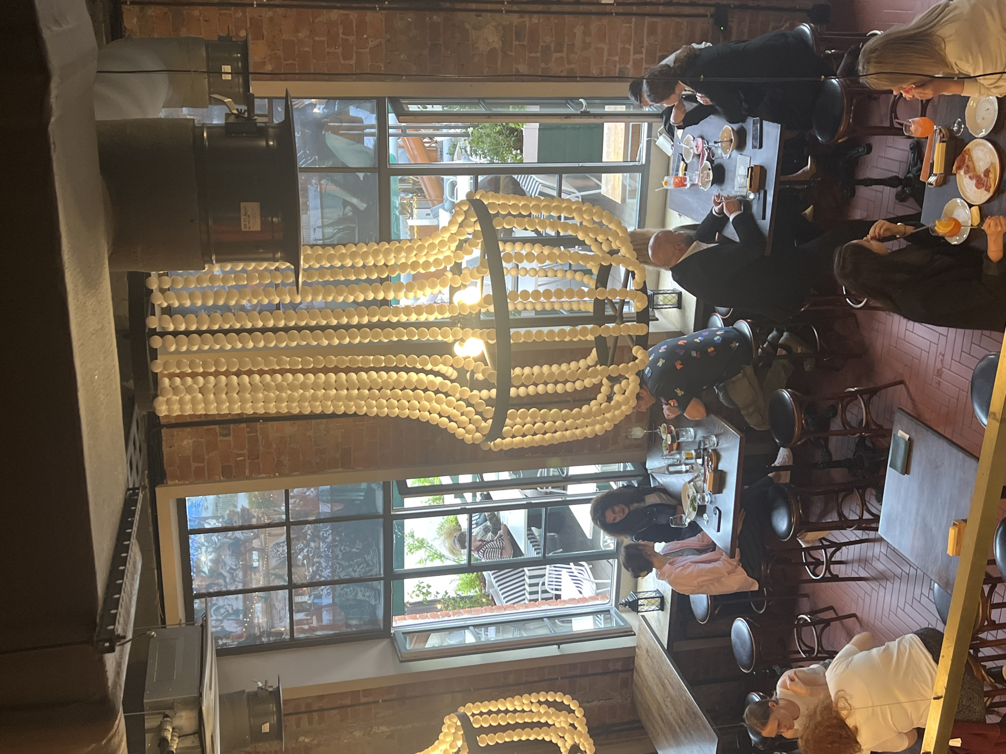 The image shows a group of people dining and socializing in what appears to be a restaurant or cafe. The setting is indoors, with a rustic brick wall and large windows offering a view of an outdoor area with greenery and more seating. A distinctive feature of the interior is a large, decorative chandelier made of numerous round, light-colored elements, likely glass or ceramic beads, suspended from the ceiling.
Several tables are occupied by diners. At one table, a group is engaged in conversation and eating, with drinks and plates of food visible. Another table has individuals seated at bar stools, some appearing to be waiting or engaged in discussion. In the foreground, a few people are seated at a table, one of whom is holding a glass.
The overall atmosphere seems casual and social. The lighting suggests it could be daytime or early evening. The presence of patrons suggests a lively environment. The architecture and decor hint at a modern establishment with industrial or bohemian influences. No specific text is legible in the image. The provided location context of Saint Petersburg, Russia, adds to the understanding of the geographical setting.