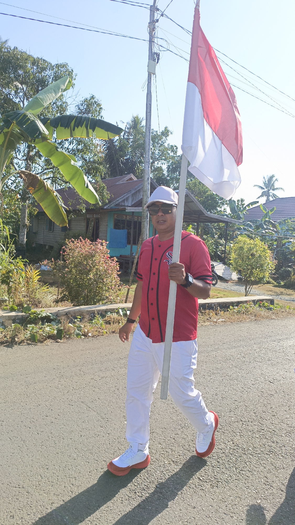 A man wearing a red baseball jersey, white cap, and sunglasses walks on an asphalt road holding a flag. The flag is red and white, striped horizontally. The man is wearing white pants and white and red sneakers. The background shows a residential area with houses, trees, and vegetation. The sky is clear and blue. The scene appears to be outdoors in a rural or suburban setting in Batarang, Indonesia, under bright sunlight, casting shadows on the road.