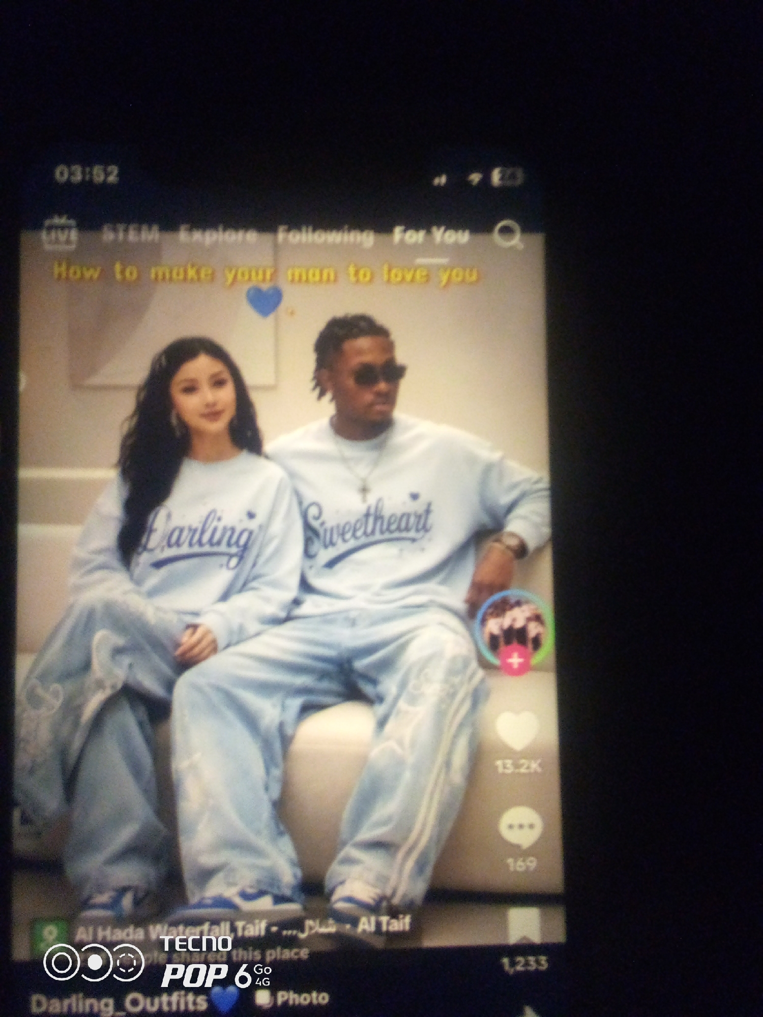 A low-angle, full-shot photograph captures a screen displaying a social media interface with a video featuring a couple. The video shows a young woman with dark, wavy hair and a young man with dark skin and dreadlocks wearing matching light blue tracksuits. The woman's tracksuit has "Darling" written in blue script on the chest, and the man's tracksuit has "Sweetheart" written in blue script. They are seated, with the man's arm resting on the back of the sofa behind them.
The screen also displays navigation elements at the top, including "STEM," "Explore," "Following," and "For You," along with a search icon. A text overlay in yellow reads "How to make your man to love you" with a blue heart icon. To the right of the video, engagement icons are visible: a heart with "13.2K," a chat bubble with "169," and what appears to be a bookmark icon with "1,233" beneath it. At the bottom of the screen, text reads "Al Hada Waterfall, Taif - ... - Al Taif" and "Darling_Outfits." A logo for "TECNO POP 6 4G" is also present. The image is a photograph of a screen, indicating the scene is likely a digitally presented one rather than a physical location. The city of Howayi, Nigeria, is not depicted or referenced in this image.