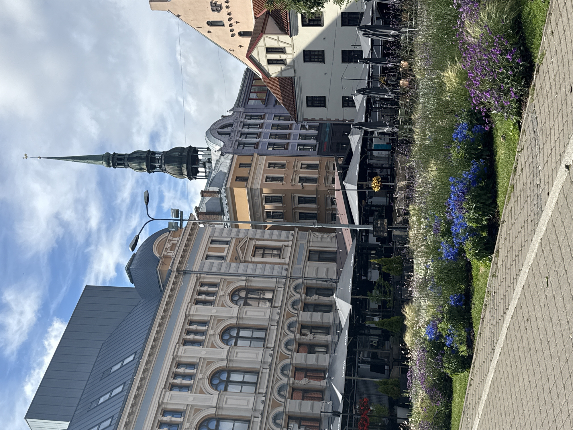 The image shows a street scene in Riga, Latvia, likely in the Old Town, with a mix of historical architecture and modern elements. In the foreground, there is a paved walkway with a decorative flowerbed to the right, featuring a variety of purple and blue flowers, along with ornamental grasses. Beyond the flowerbed, there are outdoor seating areas for cafes and restaurants, covered by large grey umbrellas. The buildings visible in the background display diverse architectural styles. One prominent building on the left has a light-colored facade with decorative elements and large arched windows, and a more modern, large grey structure behind it. To the right of this building, a tall, dark green spire with a weather vane tops a building, characteristic of St. Peter's Church in Riga. Other buildings include a pale pink structure with a triangular roof and a lighter yellow building. The sky is partly cloudy with patches of blue. No people are clearly visible or identifiable in the scene, suggesting the photo was taken during a quiet moment or the subjects are too distant or obscured. The scene appears to be a well-maintained urban environment during daylight hours.
