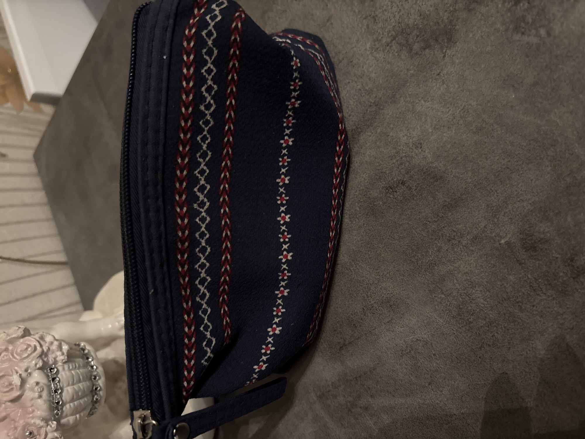 A dark blue zippered pouch, adorned with multi-colored geometric and floral patterns, rests on a textured, mottled grey-brown surface. The pouch exhibits a series of horizontal patterned bands: a top band of alternating red and white chevron weaving, followed by a band of white diamond-shaped motifs, another red and white chevron band, and a lower band featuring small white asterisk or star-like patterns, some with red centers. A metallic zipper pull and a short, flat, dark blue fabric strap are visible on the left side of the pouch. In the upper left background, a portion of a white, ornate decorative figurine, possibly ceramic, is visible, featuring a sculpted, basket-like element embellished with multiple small, reflective, gem-like beads. The upper right background contains only an indistinct, out-of-focus light surface and a warm-toned blur. The precise location depicted is indeterminable from the image data, but as requested, the scene is specified as being within the city of Chernogorsk, Russia.