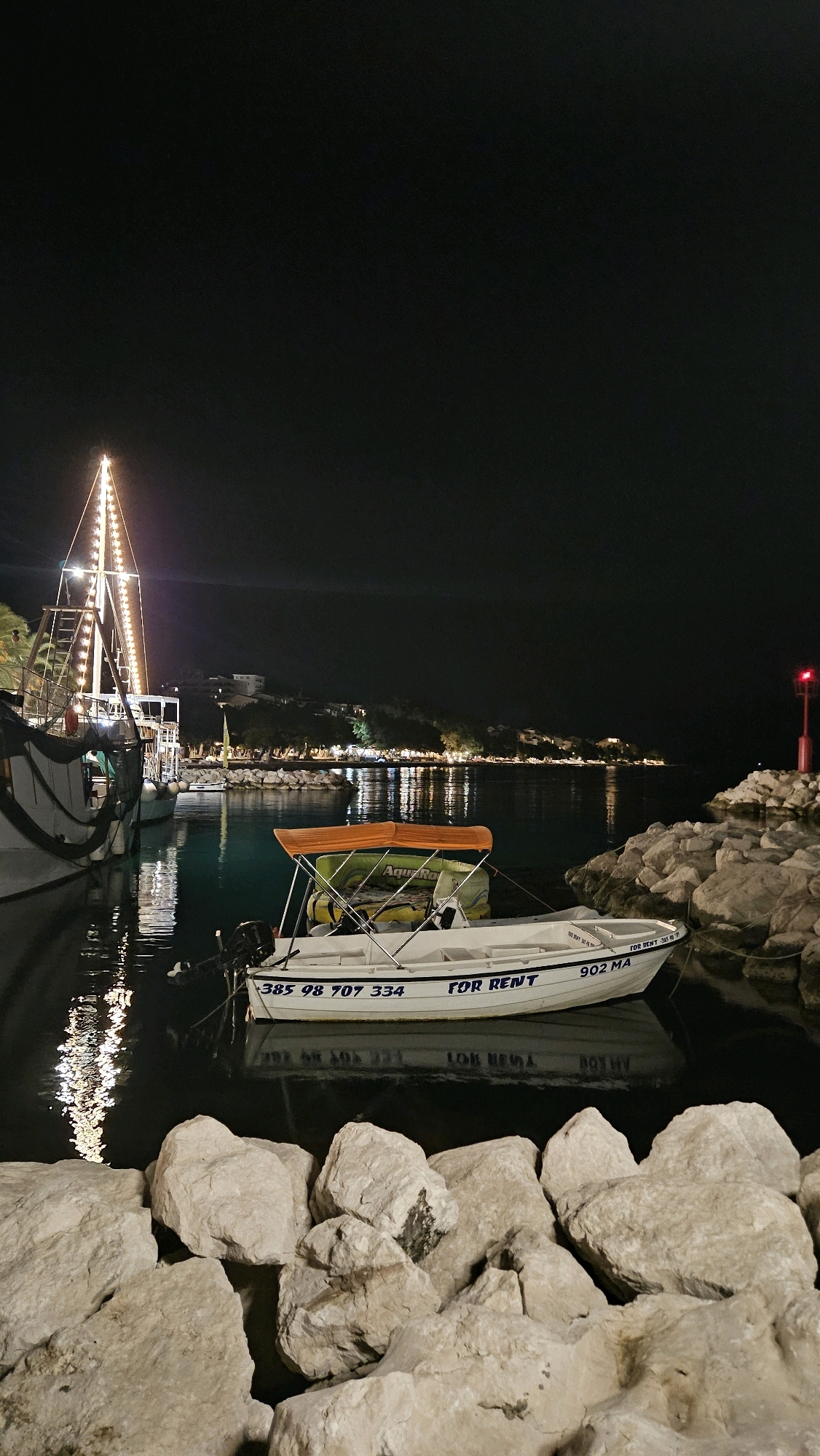 The image depicts a nighttime scene at a harbor in Općina Tučepi, Croatia. In the foreground, a cluster of large, light-colored rocks forms a barrier. Beyond the rocks, a small white motorboat is moored on dark, calm water, with its reflection visible. The boat has an orange canopy and the words "FOR RENT" are visible on its side, along with a phone number "385 98 707 334" and the designation "902 MA". In the background, a larger boat with a tall, illuminated mast is docked to the left. Across the water, the coastline is illuminated by numerous lights from buildings and streetlights, set against a dark, clear night sky. A red navigational buoy is visible to the right of the frame, marking the edge of the harbor. The overall atmosphere is peaceful and serene.