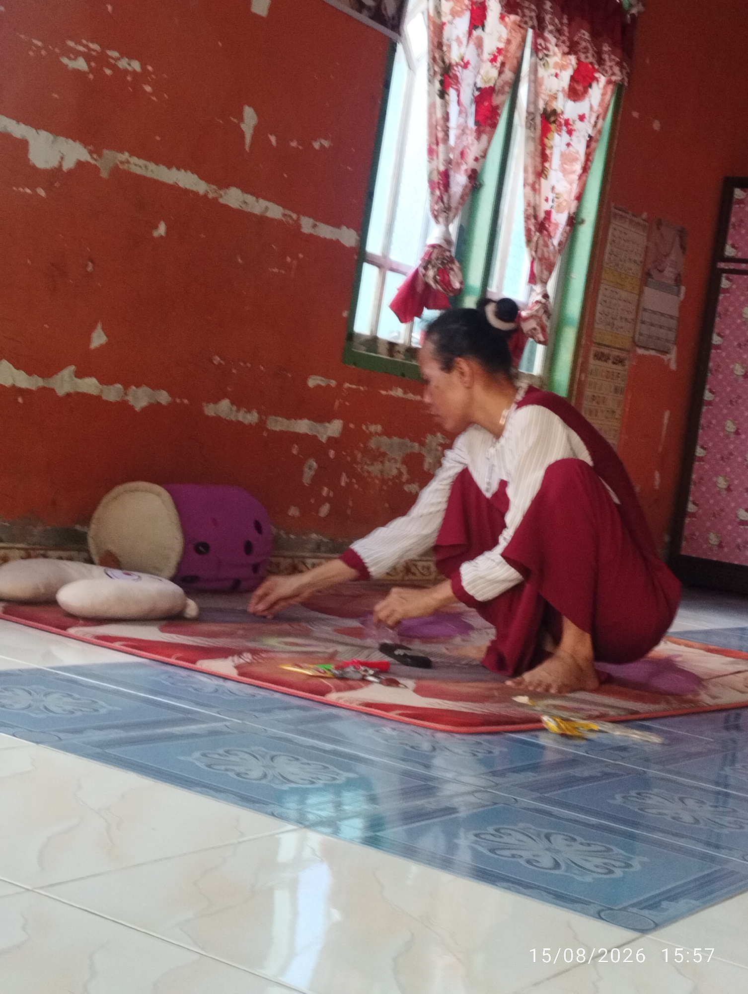 A woman, possibly Indonesian given the timestamp and location of Tegal Rejo, Indonesia, is crouching on a mat on the floor. She is wearing a white striped long-sleeved shirt and dark red pants. Her hair is pulled back in a bun. Her right hand is extended towards some items on the mat, which appear to be crafts or art supplies, including what looks like scissors and colored pencils. In front of her are several plush toys: a cream-colored round toy, a purple stuffed toy with black dots, and another cream-colored heart-shaped toy. The environment appears to be indoors, with an orange wall showing signs of peeling paint and a window with floral curtains. The floor is tiled with a patterned blue and white design. A timestamp in the lower right corner indicates the image was taken on 15/08/2026 at 15:57.
