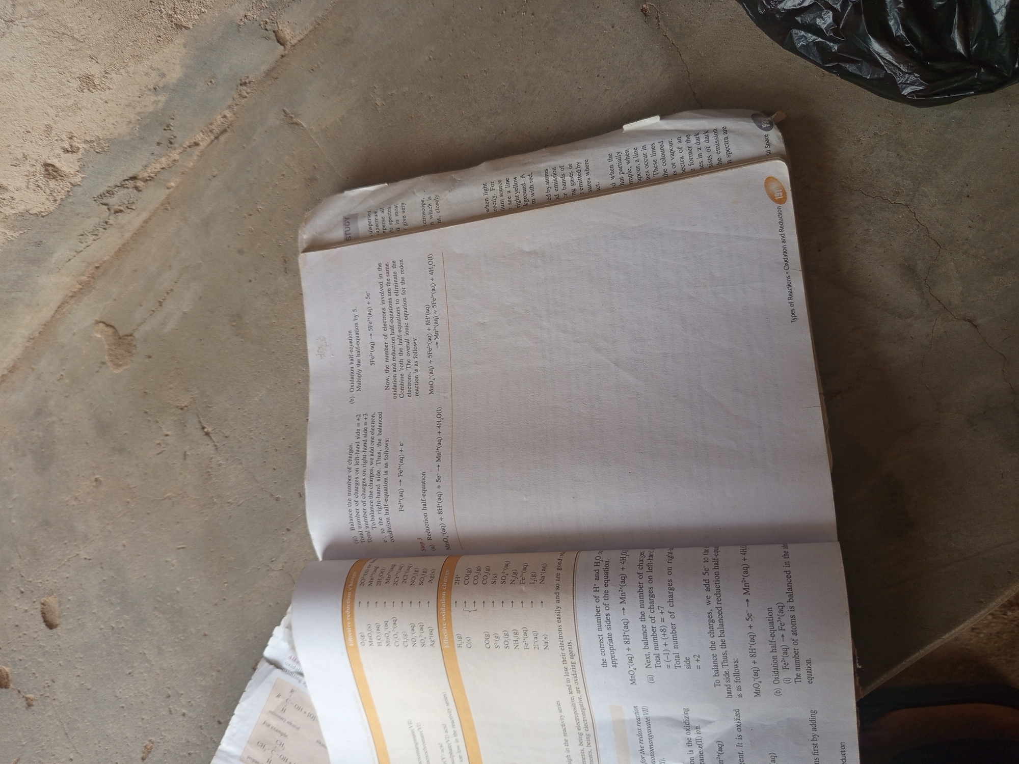 The image shows an open textbook lying on a textured, light-colored concrete surface, possibly a floor or ground. The textbook is open to a page discussing chemical reactions, specifically oxidation and reduction. The left page contains a table of elements and their corresponding effective reduction and oxidation changes. The right page has text explaining the balancing of redox reactions, with examples of half-equations and a balanced overall equation. A portion of a black plastic bag is visible in the lower right corner of the image. The overall scene is dimly lit, and there are no people present. The provided location information of Jalingo, Nigeria, cannot be verified from the image itself as there are no discernible landmarks or clues related to that specific location.