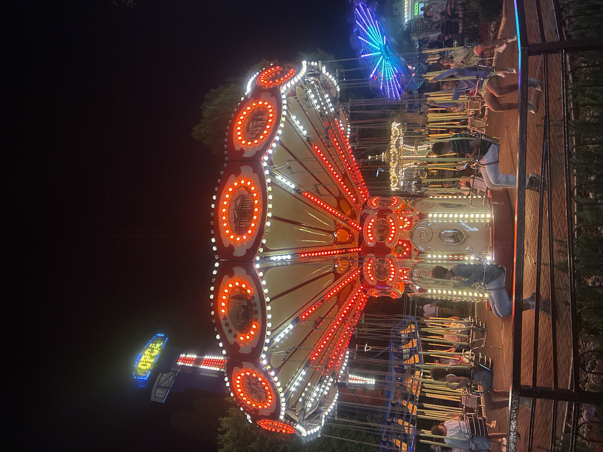 An amusement park at night, possibly in Sochi, Russia, is depicted in this image. The central focus is a brightly lit swing ride, adorned with red and white lights, in motion. Several people are seated in the swings, their forms silhouetted against the lights. In the background, other park attractions are visible, including a smaller carousel and a vibrant, multi-colored light display. Trees frame the scene, suggesting an outdoor setting. A sign with blue and yellow lettering, possibly indicating the park's status or name, is visible in the upper left corner. The ground is paved with brick, and a dark metal railing runs along the foreground, separating the viewer from the ride. The overall atmosphere is one of festive activity and nighttime entertainment.