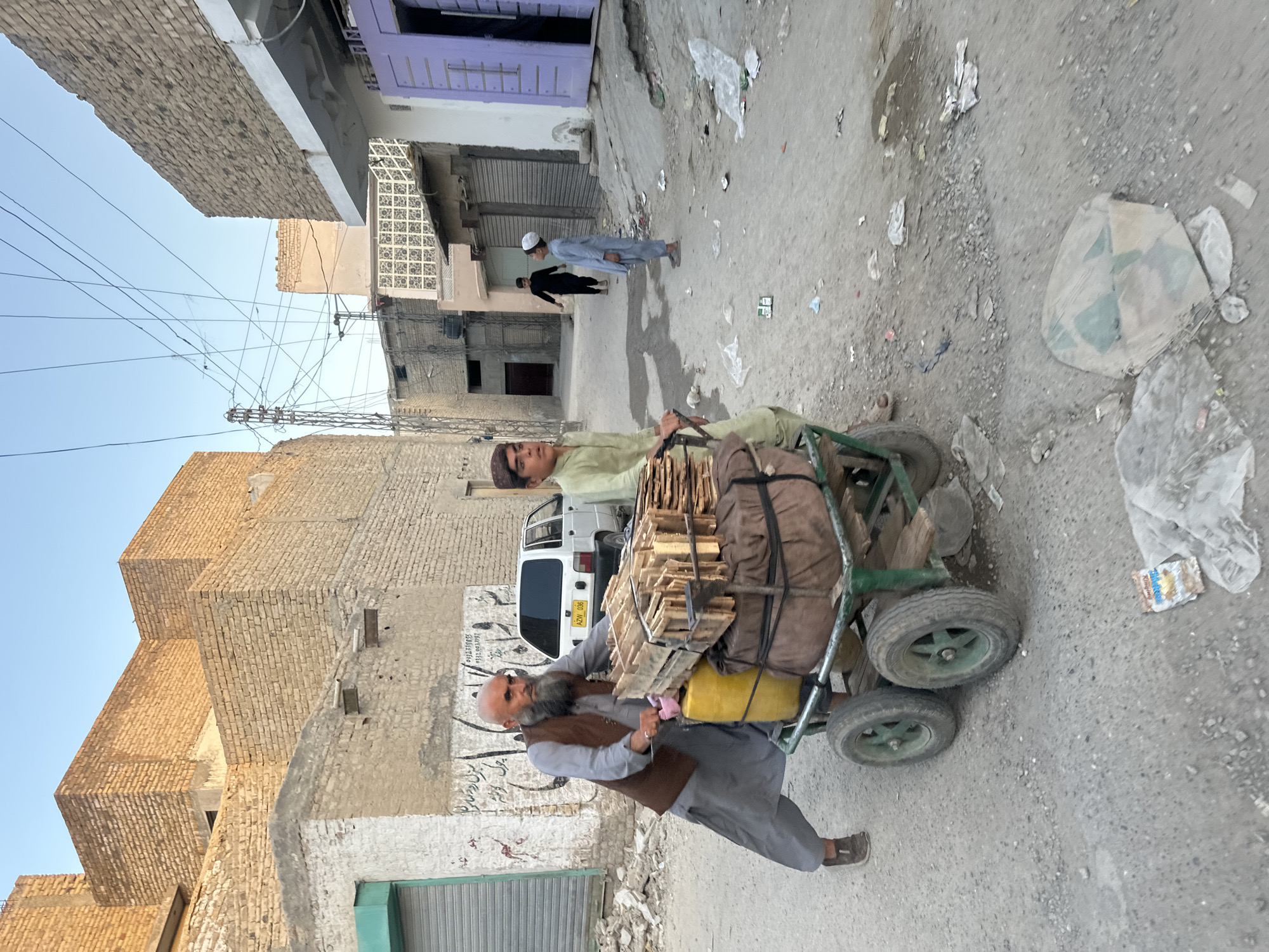 A street scene in Quetta City Tehsil, Pakistan, depicts two individuals pushing a cart laden with goods. The cart, constructed from wood and metal, carries a large yellow plastic container and stacks of wooden crates. The primary pusher, an older man with a beard and wearing a traditional shalwar kameez with a vest, is actively engaged in moving the cart. Beside him, a younger individual, also in shalwar kameez, assists. The street is unpaved and littered with debris. In the background, several buildings constructed of brick are visible, along with a white car parked on the side of the street. A power pole and electrical wires are present, indicating urban infrastructure. The sky is clear, suggesting daylight conditions.