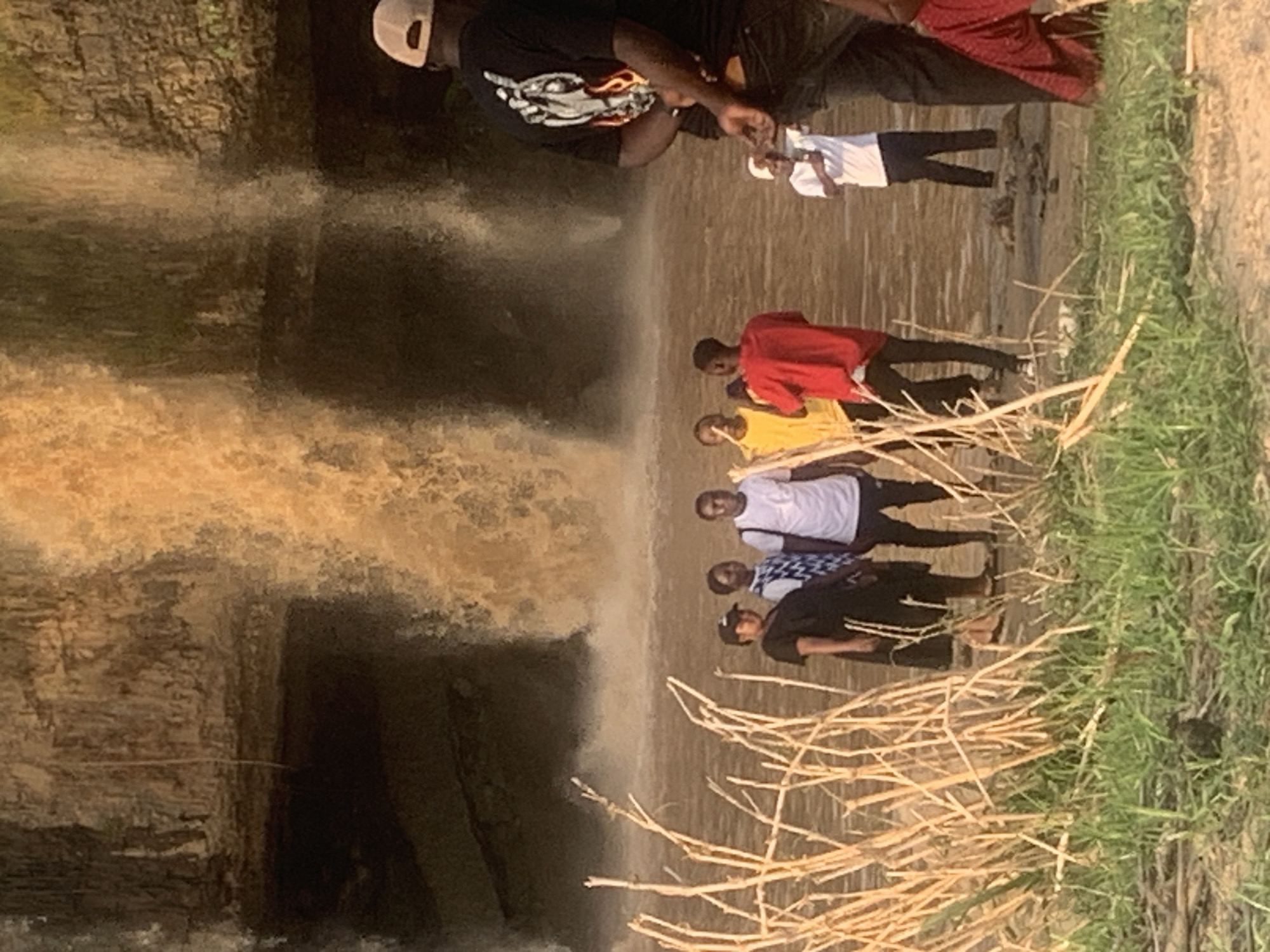 The image shows a group of people at Gurara Falls in Kukum, Nigeria. Six people are standing near the edge of the waterfall. One person is taking a picture with a mobile device, while another person is holding the mobile device being used. The group stands on land near the waterfall, which is flowing heavily. The environment is natural, with vegetation along the edge and the rocky terrain of the waterfall visible.