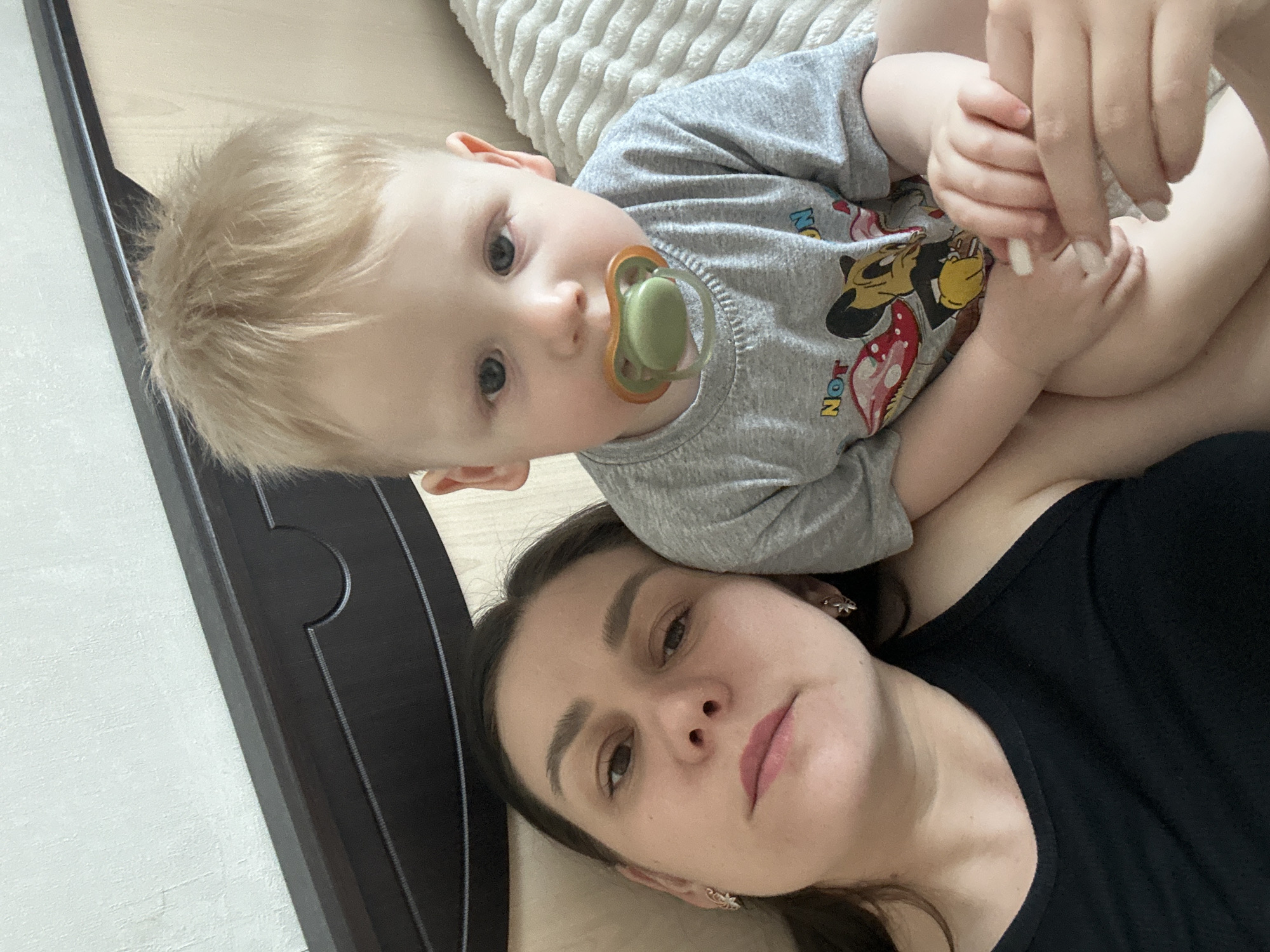 A woman lies on her back on a beige surface, potentially a bed or floor, with a young child lying partially on top of her. The child, who has blonde hair and a pacifier in his mouth, is wearing a gray t-shirt with a cartoon character on it. The woman has dark hair and is wearing a black top. The photo appears to be taken indoors in a domestic setting, possibly in Chaikovsky, Russia, as per the prompt. The child is holding and playing with someone's hand.