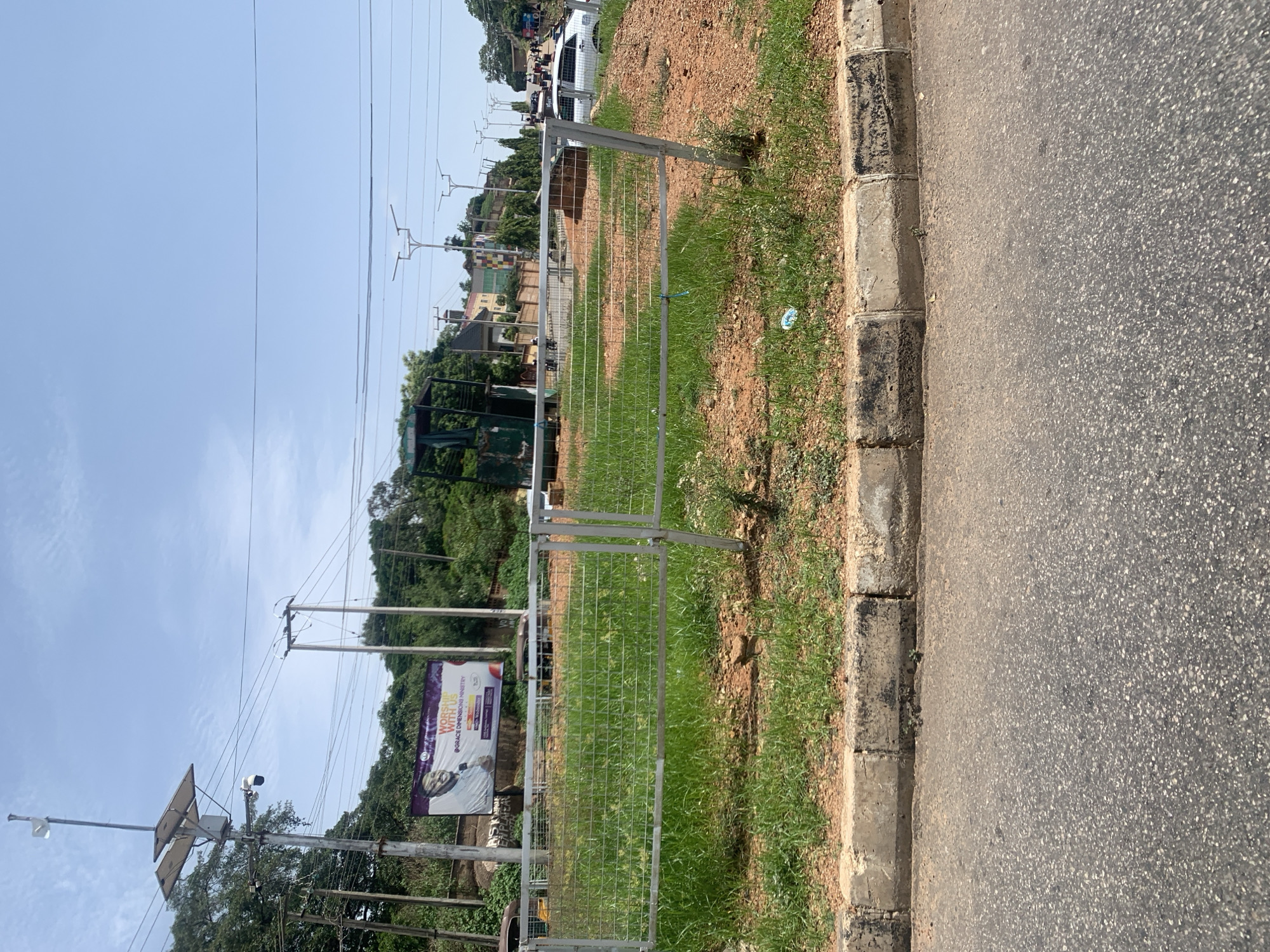 The image depicts a street scene in Jos, Nigeria. In the foreground, a grey asphalt road is bordered by a concrete curb with patches of dirt and sparse grass. A low, grey metal fence encloses an area of green grass and reddish-brown dirt. In the background, a billboard with a smiling man and text is visible, positioned behind the fence. Several utility poles with wires and solar panels are scattered throughout the scene. A large green structure, possibly a generator or a piece of equipment, stands near the billboard. In the distance, a white car is parked on the right side of the road, and other vehicles and buildings are visible, suggesting an urban environment. The sky is clear and blue with some clouds.