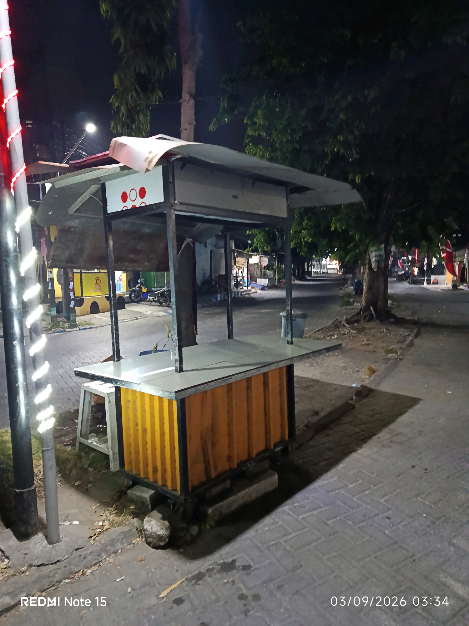 An empty food stall stands on a sidewalk at night in Keboansikep, Indonesia. The stall has a white, curved roof and an orange corrugated metal base. The sidewalk is paved with grey bricks. A pole with white and red lights is to the left of the stall. In the background, trees line a street with other stalls and parked motorcycles. The overall scene is dimly lit, indicating it is likely late at night or early morning.