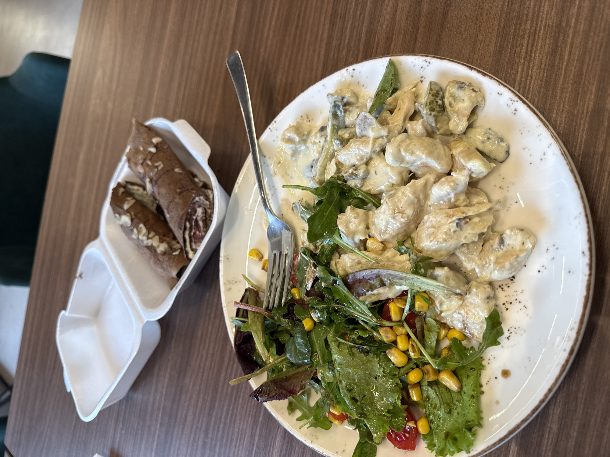 A meal is presented on a light brown wooden table in a casual dining establishment in Sofia, Bulgaria. In the foreground, a white, round plate holds a main course and salad. The salad, occupying the left side of the plate, consists of mixed green leaves including lettuce and arugula, garnished with corn kernels and halved cherry tomatoes, dressed with a clear liquid. A silver fork rests diagonally across the salad. The right side of the plate features pieces of meat, likely chicken, in a creamy white sauce with some darker inclusions, possibly mushrooms or sliced gherkins, all lightly seasoned with black specks. In the background, two white styrofoam takeout containers are visible. The front container holds two chocolate-colored crepes, rolled and filled with a lighter cream, topped with slivered almonds. The crepe on the left is partially cut open, revealing its filling. The second container behind it appears empty. The background shows a blurry dark green upholstered seating area.