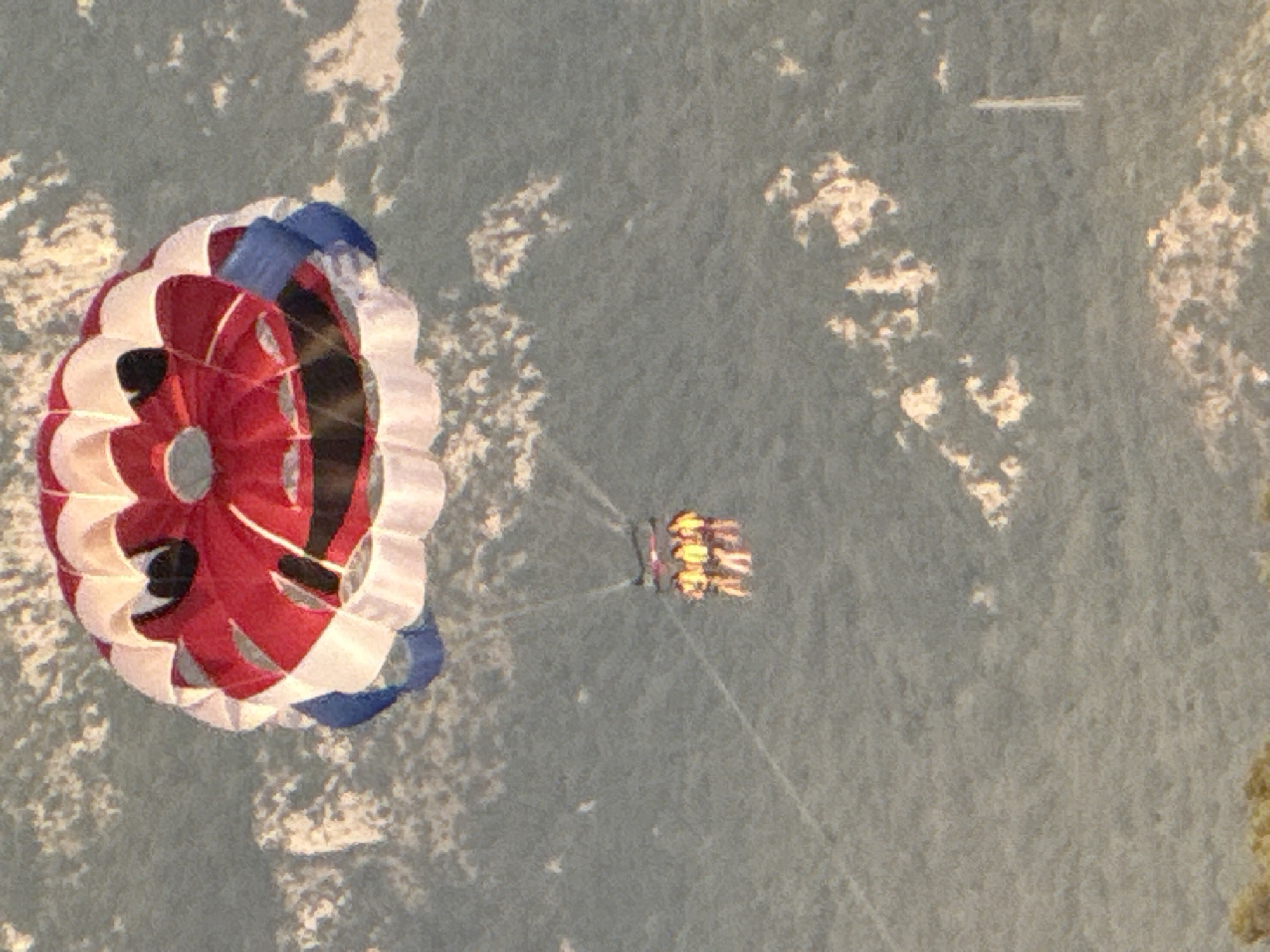 The image shows a parasail in the shape of a smiley face. The parasail is red and white with blue accents. Three people are visible in the harness, wearing yellow life vests. They are being towed by a boat, which is not visible in the frame. The parasail is flying over a densely forested area, which appears to be a hillside. The sky is overcast. The scene suggests a recreational activity, likely parasailing, taking place on a sunny day. The location is likely the coast of Budva, Montenegro, given the lush vegetation and hilly terrain.