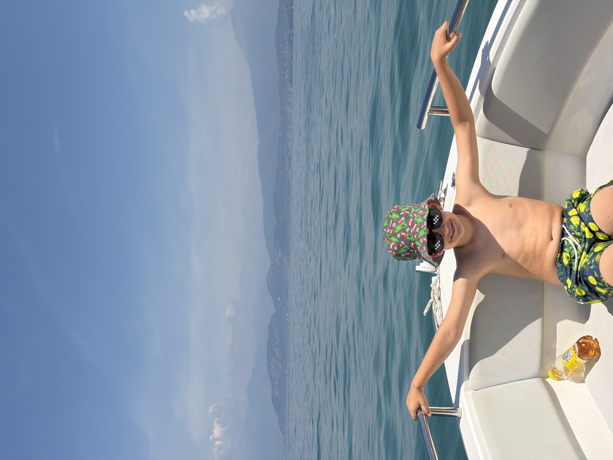 A young boy with a bucket hat and sunglasses sits in a white boat on a body of water, likely Lake Garda, near Castelnuovo del Garda, Italy. The boy is shirtless and wearing patterned swim trunks, with his arms spread out on the sides of the boat. A bottle of amber liquid is placed on the seat next to him. In the background, hazy mountains are visible across the water under a clear blue sky.