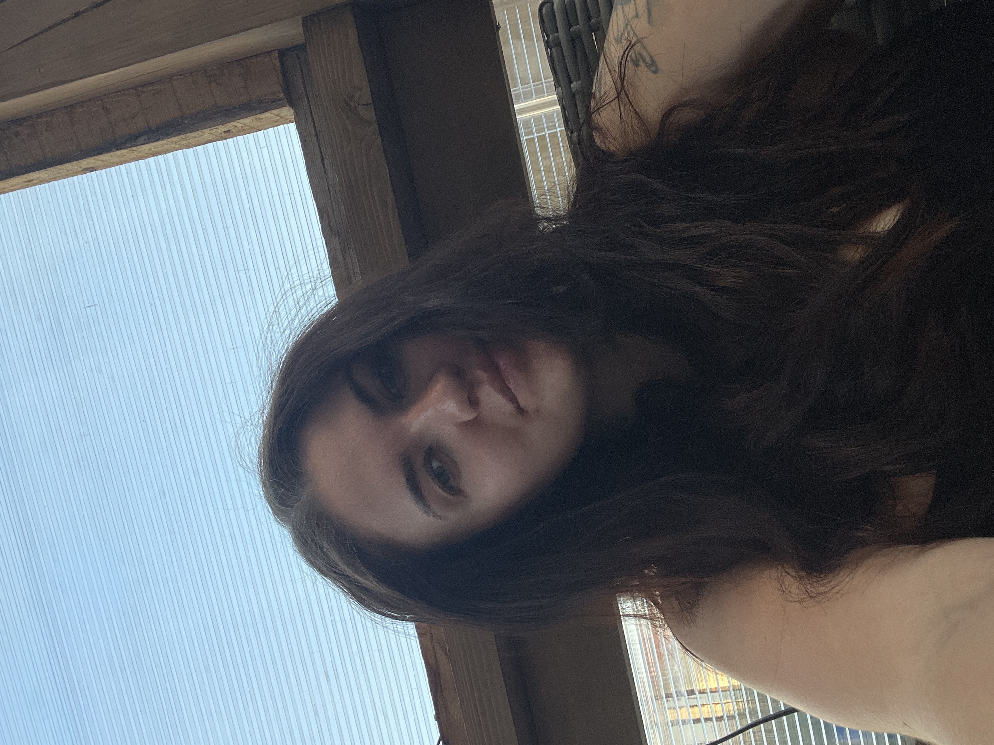 A low-angle shot shows a woman with long, dark hair. Her face is visible, and she appears to be looking down at the camera. A portion of her shoulder is also visible. The background includes what appears to be a translucent corrugated plastic sheet and some wooden beams. A building is partially visible behind the translucent material. The location is Мосметаллоконструкция Онт, Russia.
