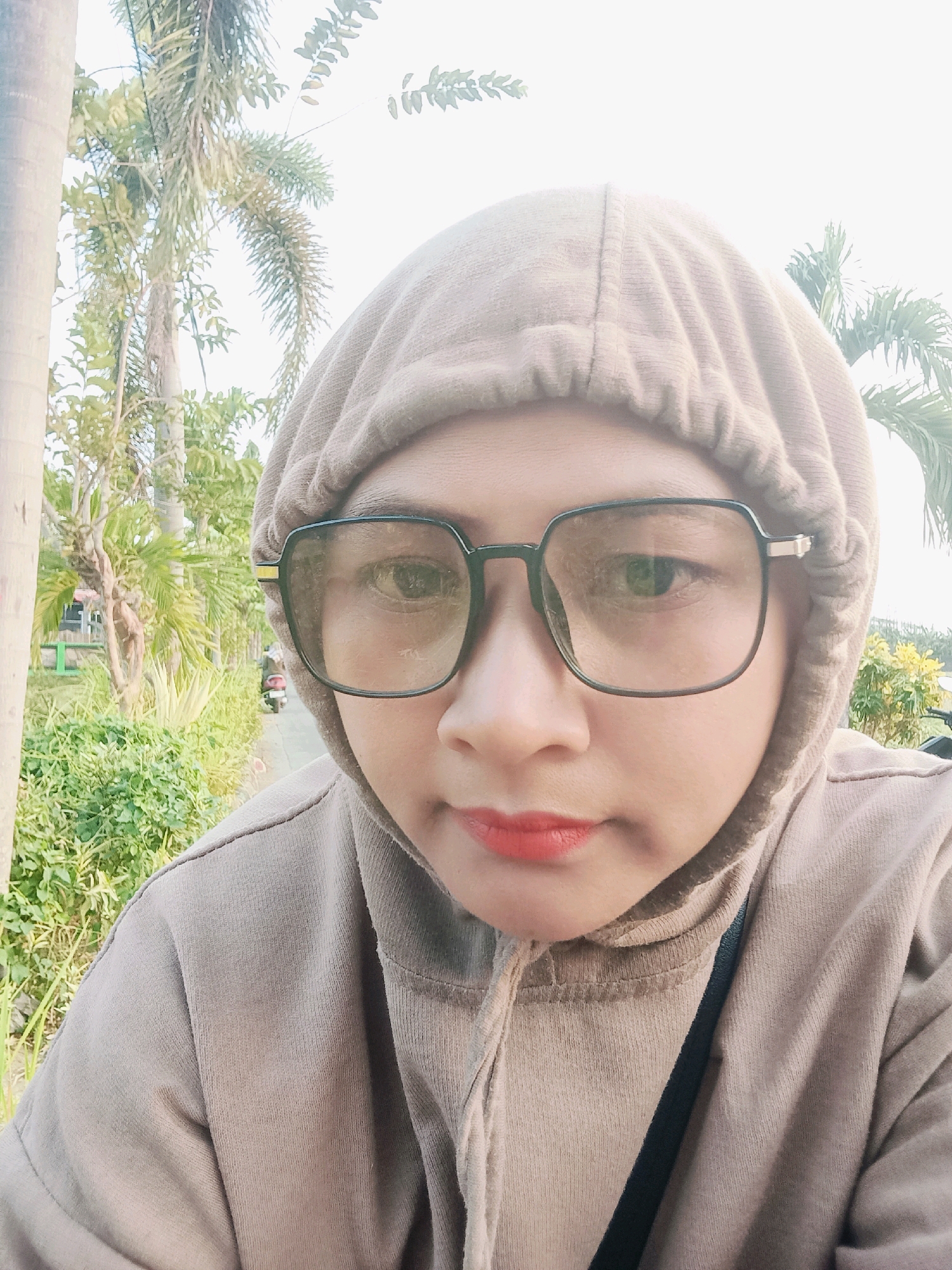 A woman wearing a beige hooded sweatshirt and large black-rimmed glasses is taking a selfie outdoors. She has red lipstick on. In the background, there are palm trees, green bushes, and a paved road with a scooter visible in the distance. The scene appears to be in a park or a residential area in Sentul, Indonesia, given the tropical vegetation. The lighting suggests it is daytime.