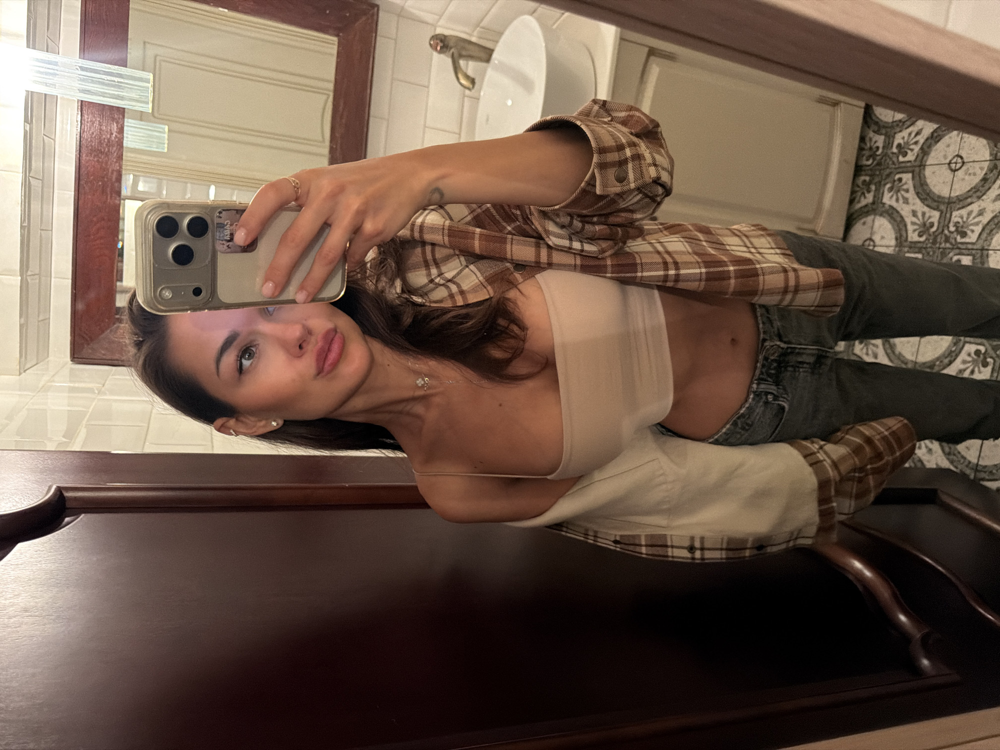 A woman is taking a selfie in a dimly lit bathroom. She is wearing a beige tube top, a plaid shirt unbuttoned and draped over her shoulders, and gray jeans. She has long dark hair and is wearing subtle makeup. She is also wearing earrings and a delicate necklace. The bathroom has white tiled walls, a wooden framed mirror, and a white sink with a golden faucet. The floor is tiled with a patterned design. A dark wooden door is visible on the left side of the image. The scene appears to be in an interior residential setting.