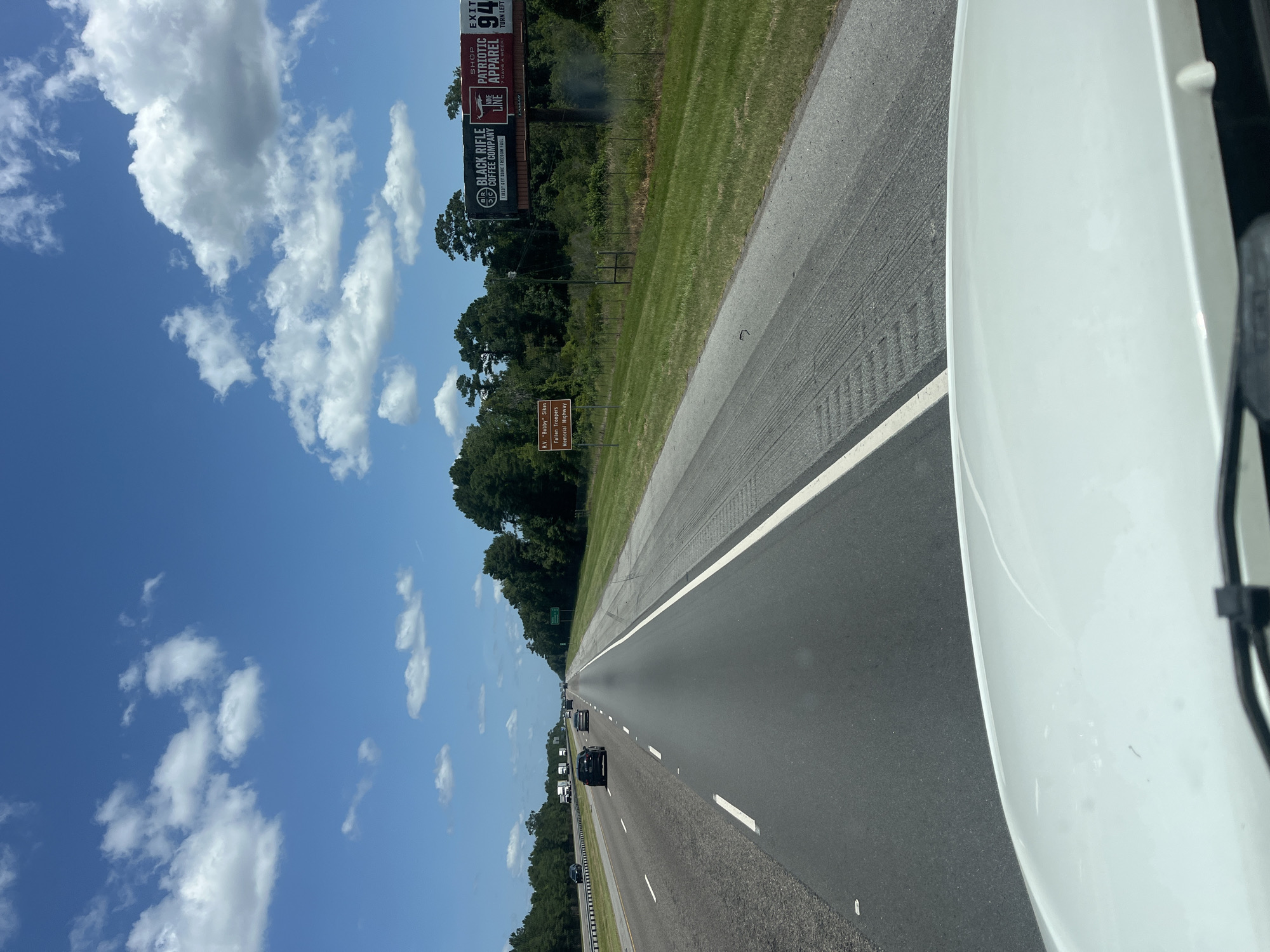 The image captures a view from the perspective of a vehicle traveling on a highway in Riceboro, United States. The road ahead is asphalt, with multiple lanes and white lane markings. Several vehicles are visible on the road, including cars and trucks. The environment features a bright blue sky with scattered white clouds. On the right side of the highway, a grassy median is present, followed by a fence and a dense line of trees. A brown sign is visible on the left side of the median, reading "R.V. 'Bobby' Sikes Fallen Troopers Memorial Highway." Further down the road on the right, a large billboard displays advertisements for "Black Rifle Coffee Company" and "Nine Line Patriotic Apparel." The foreground shows the white hood of the vehicle from which the image is taken, with a portion of the windshield wipers visible. The lighting suggests it is daytime, with strong sunlight casting shadows.