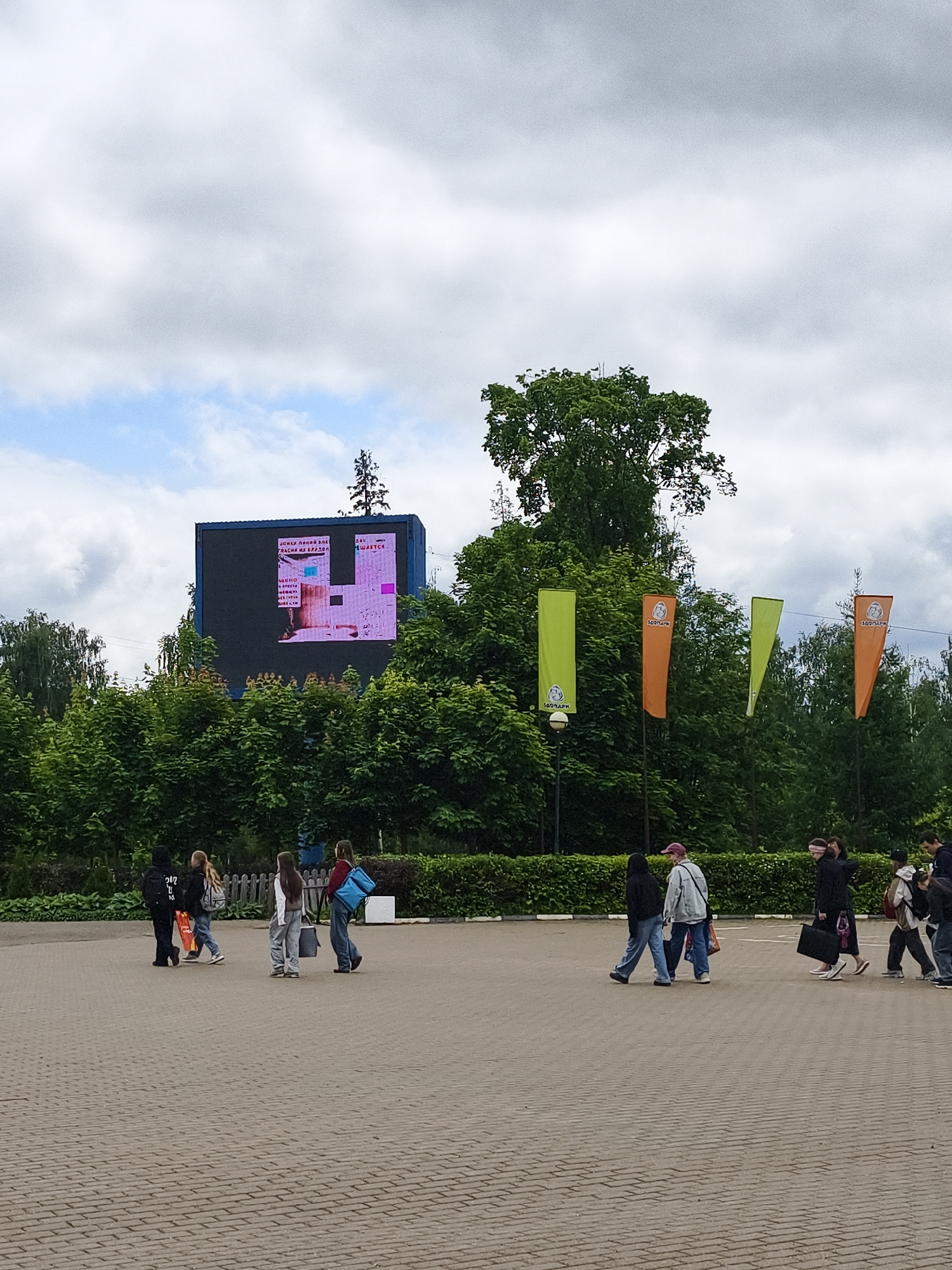An outdoor, eye-level photograph captures a group of people walking across a paved area under an overcast sky in Izhevsk, Russia. The foreground features a light-colored brick-patterned pavement.
In the midground and background, dense green foliage, comprising tall trees and bushes, lines the perimeter of the area. A prominent feature is a large, dark-framed digital screen mounted above the treeline on the left. The screen displays a colorful, pixelated image with some visible Russian text. Partial words such as "ЗАР ЛЕНИЙ ЭЛЕ..." and "...ЕШАЕТСЯ" are seen at the top, while "ЭТО" and "БИЛЕТ" are visible elsewhere on the screen, hinting at information or possibly a game/interactive display, perhaps related to event tickets.
To the right of the screen, several vertical banners, alternately colored green and orange, are hoisted on poles among the trees. Each banner prominently features a logo containing the Russian word "ЗООПАРК" (ZOOPARK), strongly indicating that the location is the Izhevsk Zoo or an entrance to it.
Approximately ten people, primarily young adults or teenagers, are seen walking across the paved area. They are dressed in casual attire including hoodies, jackets, jeans, and t-shirts, with some carrying backpacks or tote bags. Their relaxed postures suggest a leisurely activity. The sky is largely covered with clouds, indicating daytime with diffused natural light. The scene depicts a casual gathering at a public outdoor venue, likely an entrance or plaza leading to the zoo.