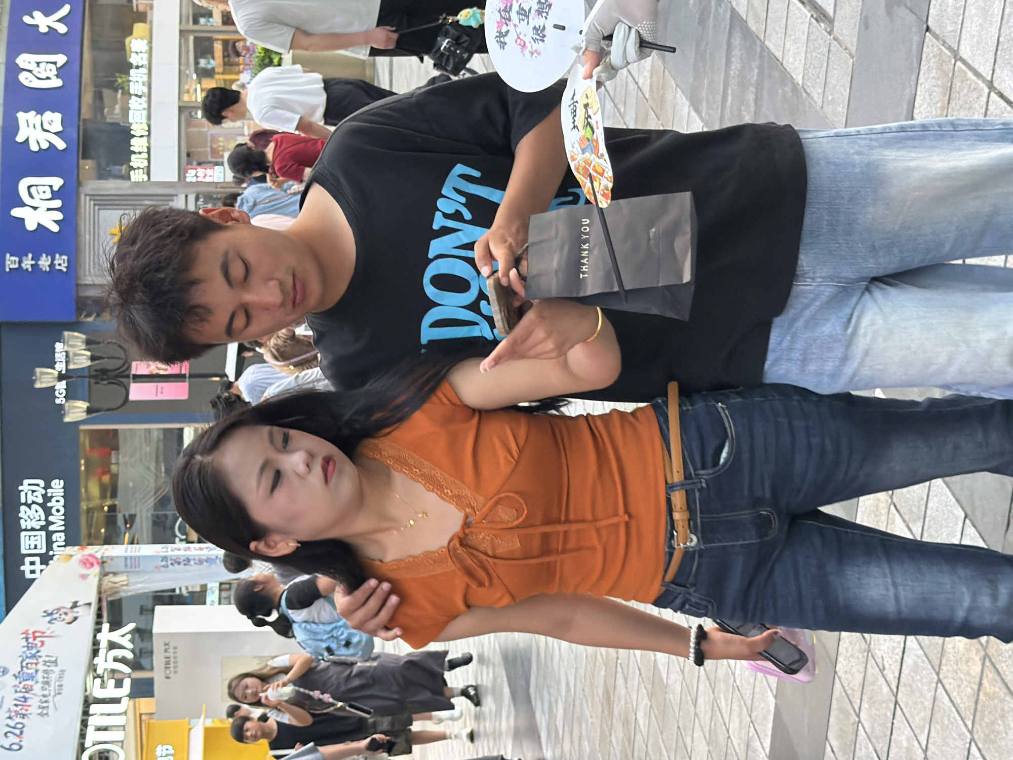 In the Yuzhong District of China, a young couple stands close together on a tiled pedestrian walkway. The man, wearing a black t-shirt with blue lettering that reads "DON'T," has his arm around the woman. He is looking down at a mobile phone in his hand, which he is holding with his left hand. The woman, in an orange short-sleeved top and blue jeans, is also looking down at the phone, her right hand resting on the man's arm. A dark gray paper bag with "THANK YOU" printed on it is held by the man. In the background, other pedestrians are visible, along with storefronts and signage in Chinese. The overall scene suggests a casual outing in a bustling urban environment.