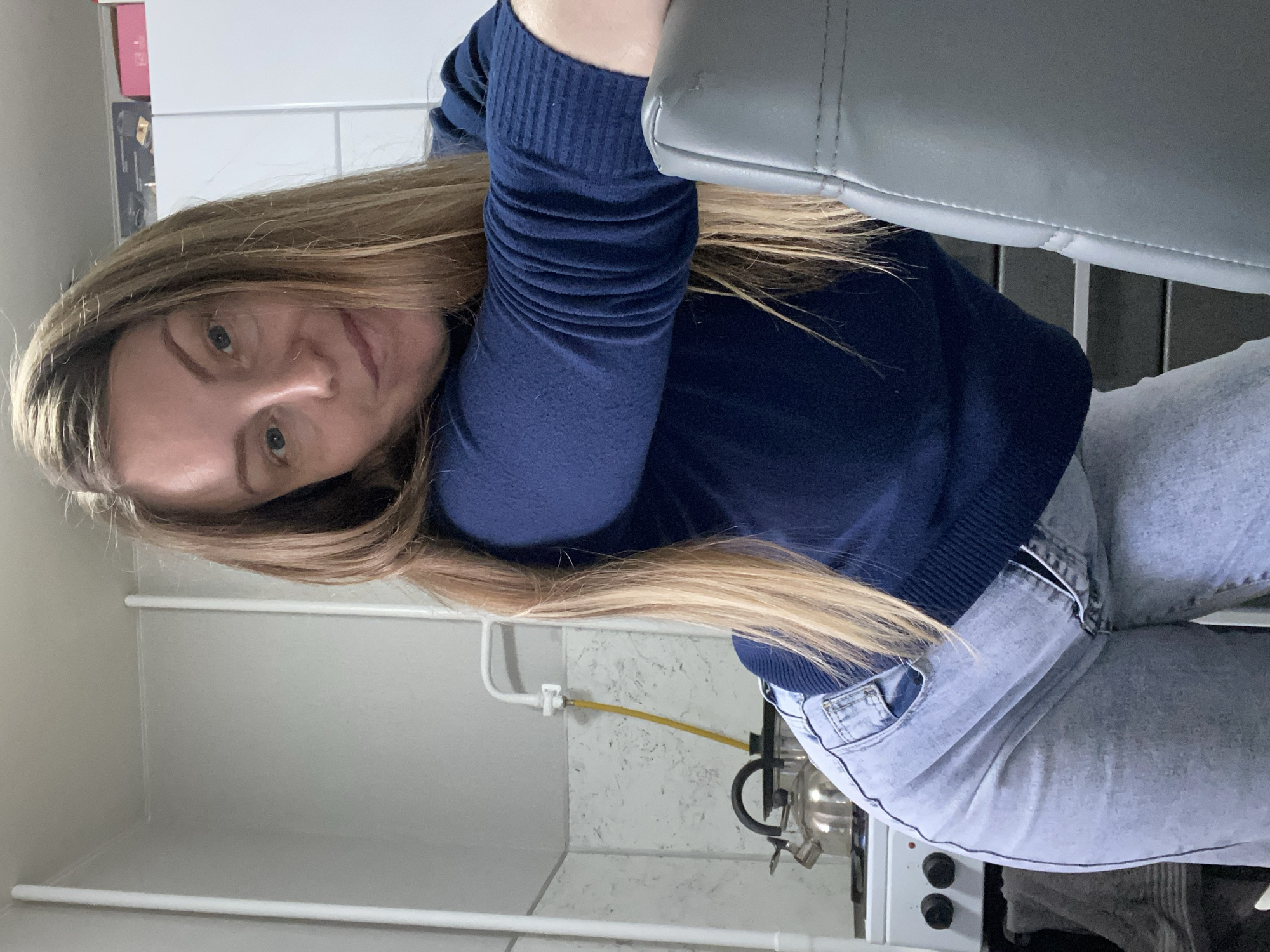 A young woman with long, straight, light brown hair is seated indoors, likely in a kitchen setting in Noviy, Russia. She is looking directly at the camera with a neutral expression, her head tilted slightly. She wears a dark blue, long-sleeved ribbed top and light blue denim jeans. Her left arm is bent at the elbow, resting on the gray, possibly leather, back of a chair.
The background reveals a kitchen area with white tiled walls and a section with a lighter, possibly textured or marble-patterned surface. A yellow flexible gas hose is visible, connected to a white fixture on the wall, leading towards a gas stove or hob. A silver-colored kettle with a black handle is positioned on the stove, which features at least two black control knobs. Below the gas connection, there are white horizontal bars, possibly part of a shelving unit or furniture. The lighting appears bright and even, suggesting it is daytime. There is no visible text in the image.