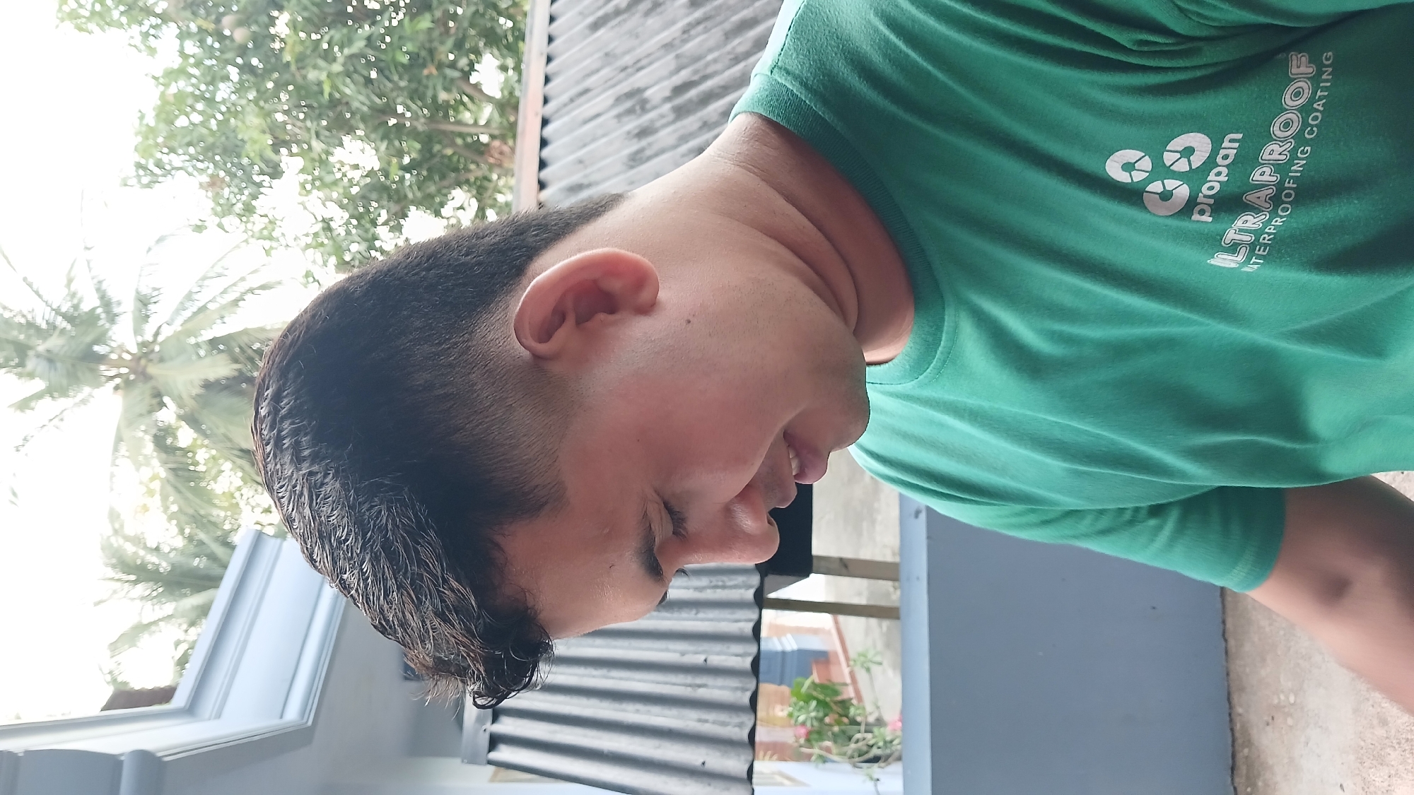A medium shot depicts a man wearing a green shirt with a company logo partially visible in the top right corner. The shirt reads "propan" with three circular shapes stacked above it, followed by "LTRAPROOF WATERPROOFING COATING" beneath. The man has short, dark hair, light skin, and his eyes are closed. His head is slightly tilted forward.
The background shows elements of a residential setting. A palm tree is visible in the upper left corner. Part of a building with white trim and a corrugated metal roof can be seen. There is also some greenery, possibly plants in pots, and a gray surface that might be a wall or patio.
The image appears to have been taken outdoors in Lhokseumawe, Indonesia.
