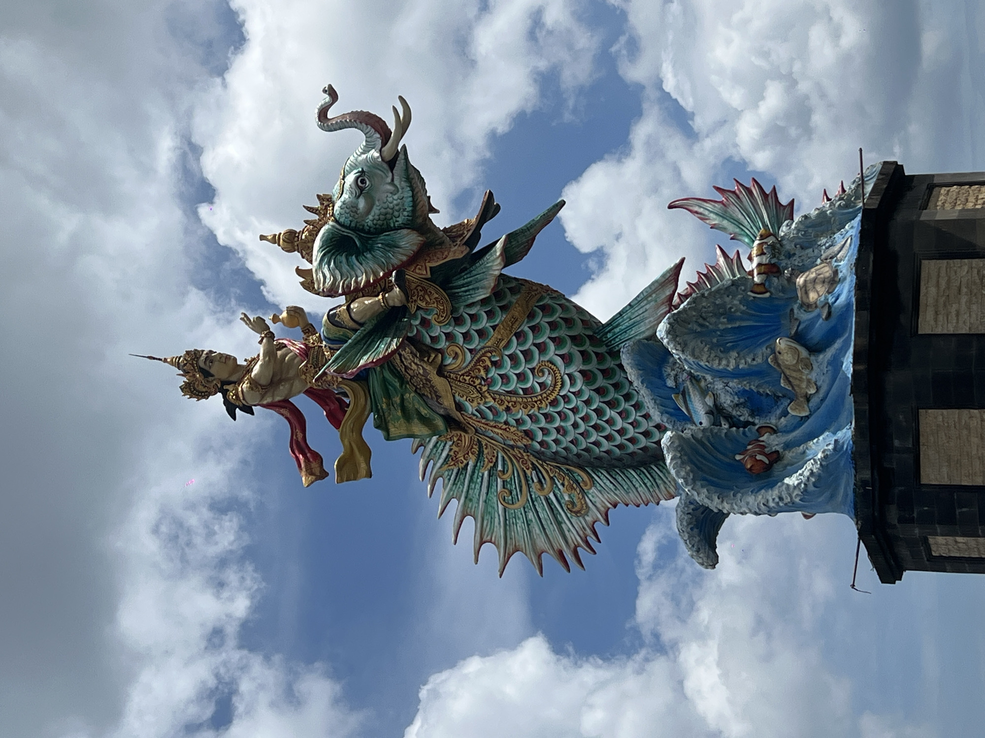 An intricately detailed, vibrant statue stands prominently against a bright, cloudy blue sky, captured in what appears to be North Kuta, Indonesia. The central subject is a majestic, mythical creature resembling a large, ornate fish or dragon. Its body is covered in iridescent green and blue scales with golden accents, leading to elaborate fins and a multi-spiked tail in shades of green and red. The creature's head is adorned with horns or antlers and features a snouted face, its eye looking forward.
Emerging from the back of this creature is a human-like figure, possibly a deity or mythical being, clad in rich, flowing garments of red, gold, and beige. This figure wears an elaborate golden headdress and holds what appears to be a small golden vessel.
To the right of the main creature, the sculpture transitions into a dynamic blue wave-like structure, depicting smaller sea creatures such as orange and white clownfish and green turtles. This wave element connects to a darker, block-like architectural structure, possibly a part of a building or temple gate. The sky above is a mix of bright blue and scattered white cumulus clouds, indicating a daytime setting. The entire sculpture is characterized by its rich colors, intricate detailing, and mythical theme, reflecting a traditional Southeast Asian artistic style. There is no visible text.