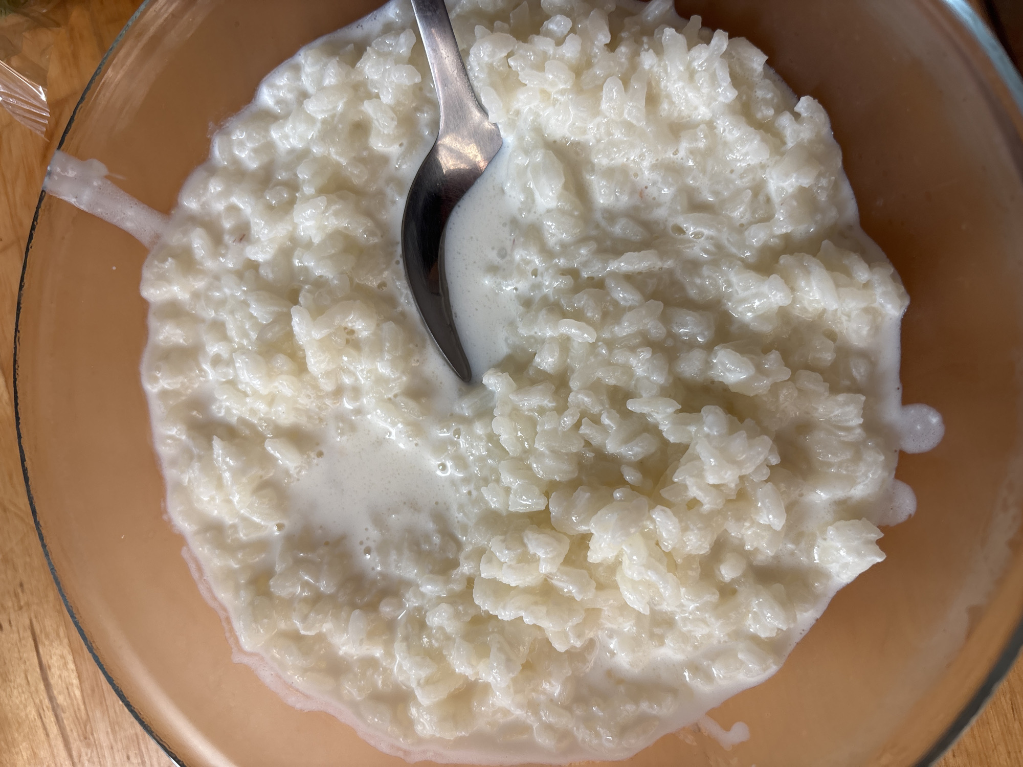 A close-up, top-down view captures a bowl of creamy rice pudding with a silver spoon partially submerged in it. The main subject is the rice pudding, which appears thick and white or off-white, with distinct, plump grains of rice visible throughout. A generous amount of milky liquid surrounds the rice, with some small bubbles present on the surface, suggesting its rich texture.
The pudding is served in a clear glass bowl, which has a slight light brown or amber tint, especially noticeable where the liquid extends to the edges. The spoon is positioned near the center-left of the bowl, its reflective surface catching some light.
The setting appears to be indoors, possibly on a wooden table, as indicated by the light brown wooden surface visible around the rim of the bowl in the background. In the upper left corner, a small, crinkled piece of translucent material, possibly plastic wrap or foil, is visible near the edge of the bowl. The image focuses entirely on the food, providing no cues about the time of day, weather, or emotions. There is no visible text.