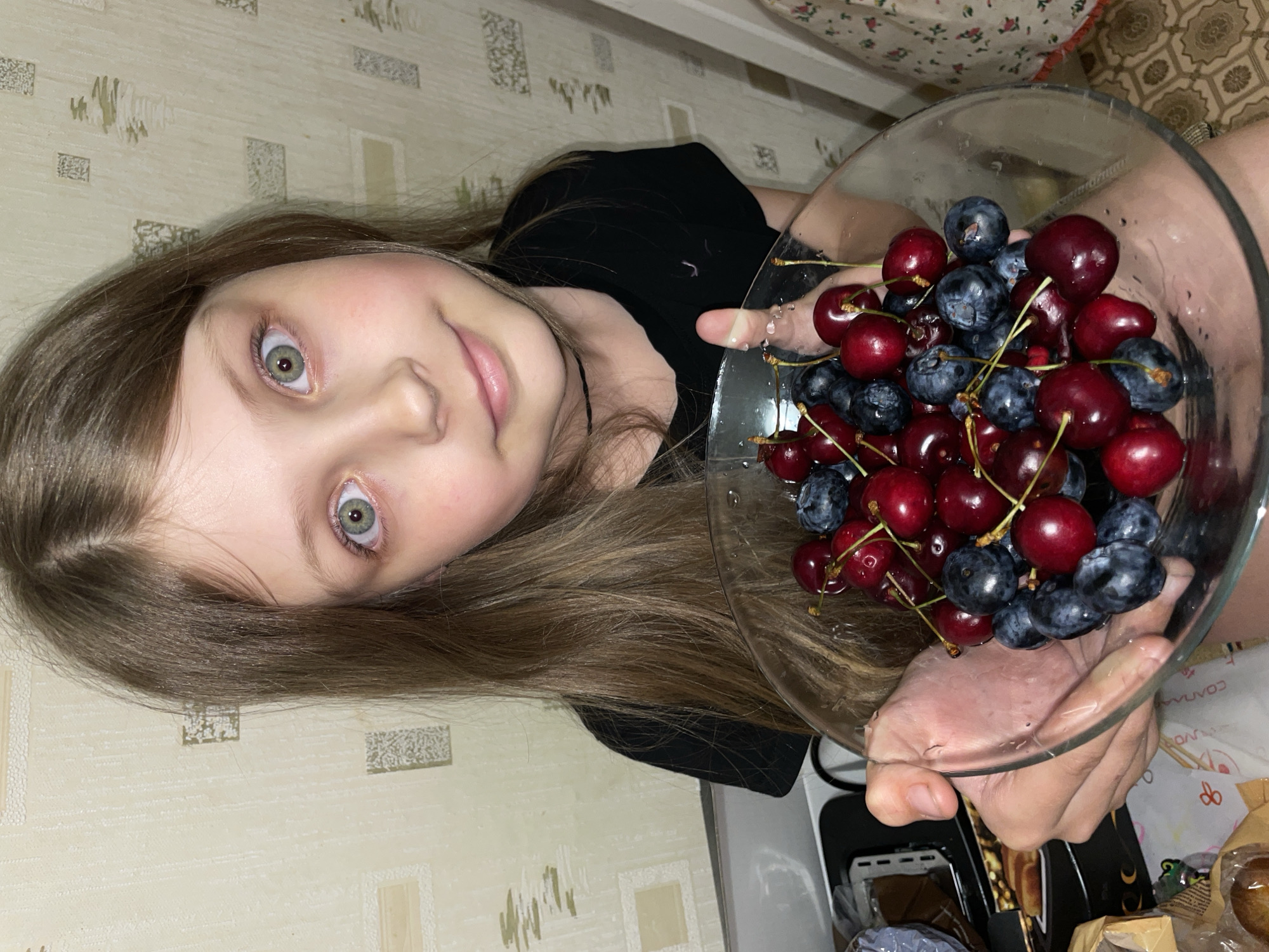 A young girl with long brown hair and green eyes is holding a clear glass bowl filled with cherries and blueberries. She is wearing a black shirt. The background shows a patterned wall and part of a kitchen counter with a white appliance. The location is unknown and cannot be determined from the image.