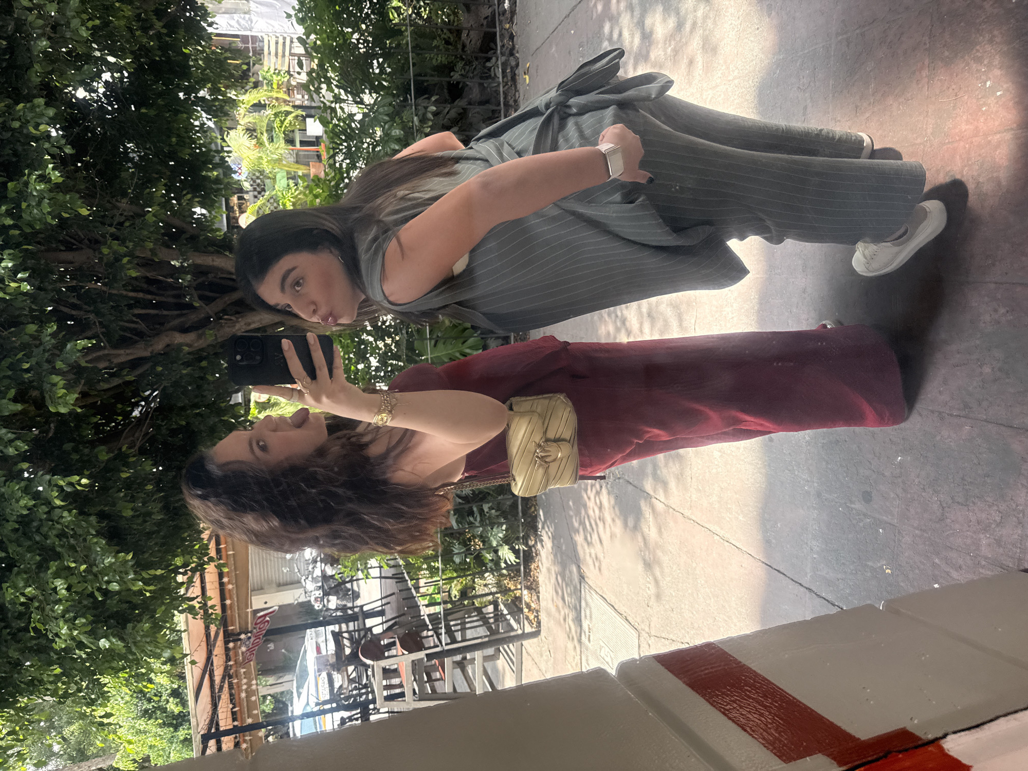 Two women are taking a selfie in what appears to be Mexico City, Mexico. One woman is wearing a red dress and a light-colored purse. The other woman is wearing a gray pinstripe jumpsuit. The women are standing on a sidewalk. Behind them are trees and a building.