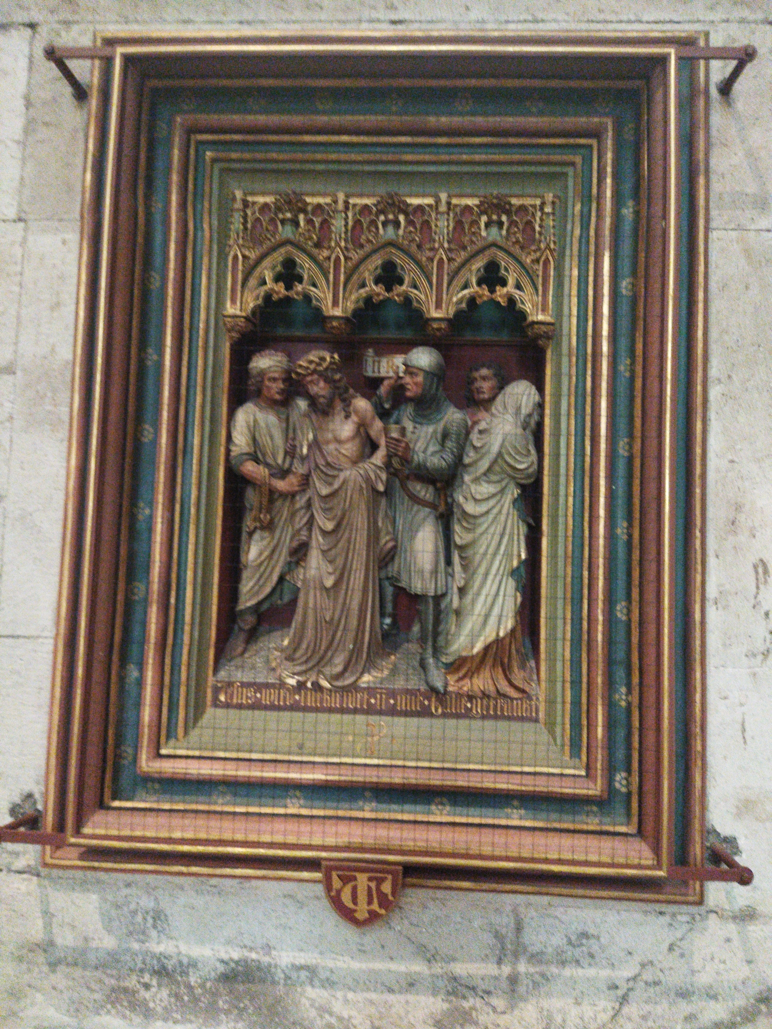 The image shows a detailed carving or relief artwork framed by an ornate, multi-layered border. The artwork itself depicts a biblical scene, likely the Passion of Christ, with four figures. On the left, a figure wearing a helmet and a robe holds a cup. Next to him, another figure, presumably Jesus, is depicted with a crown of thorns and a draped garment. To the right, two other figures are present, one wearing a hooded robe and the other a flowing garment. The figures are rendered in a realistic style. The artwork is housed within a decorative frame that features Gothic architectural elements like pointed arches and intricate carvings. The entire piece is mounted on a stone wall. A small red shield with an emblem is visible below the frame. This artwork is located in the St. Gereon Church in Monheim am Rhein, Germany.