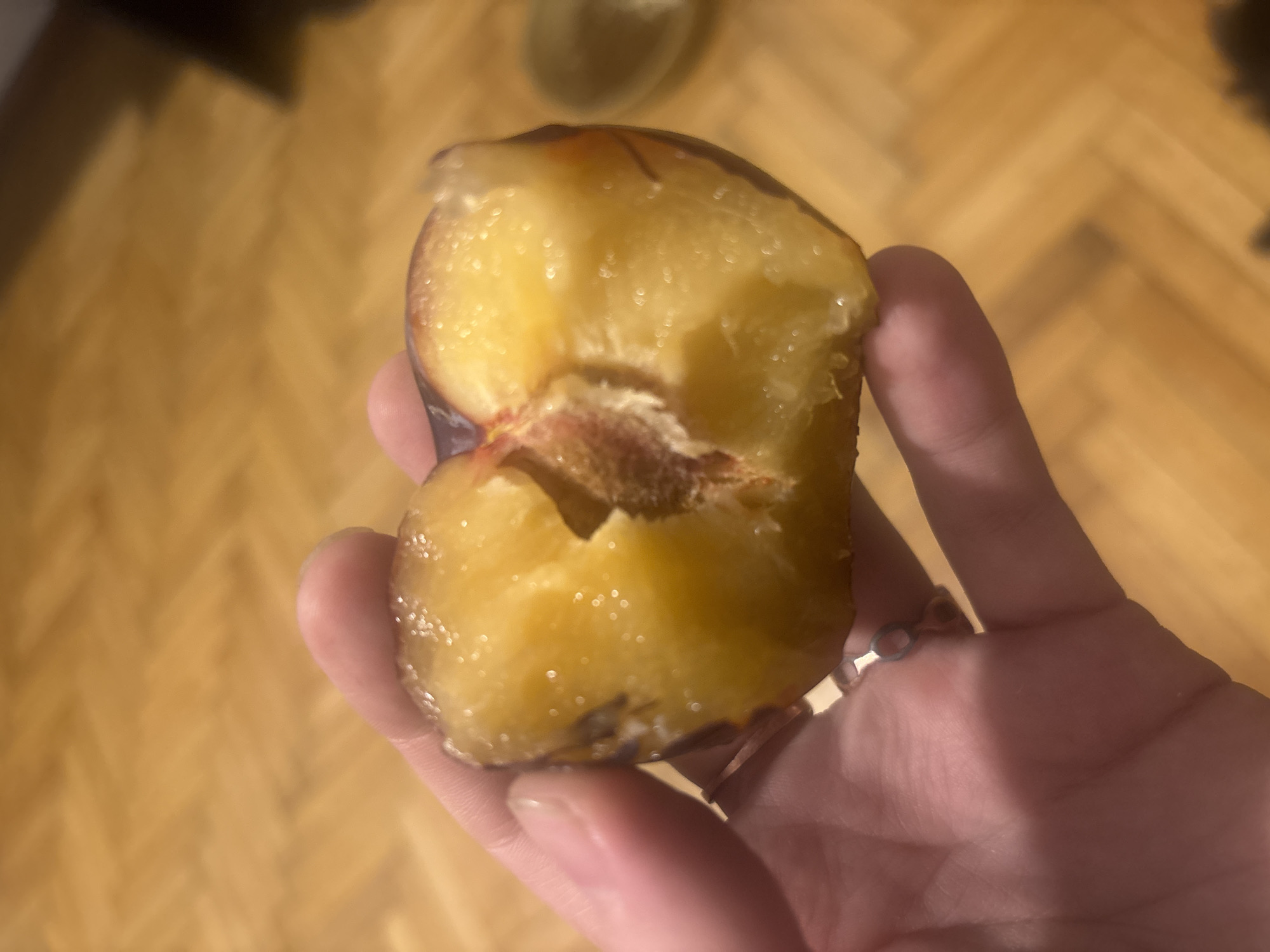 A human hand holds a halved plum. The plum has yellow flesh and a reddish-brown pit. The background is a wooden floor.