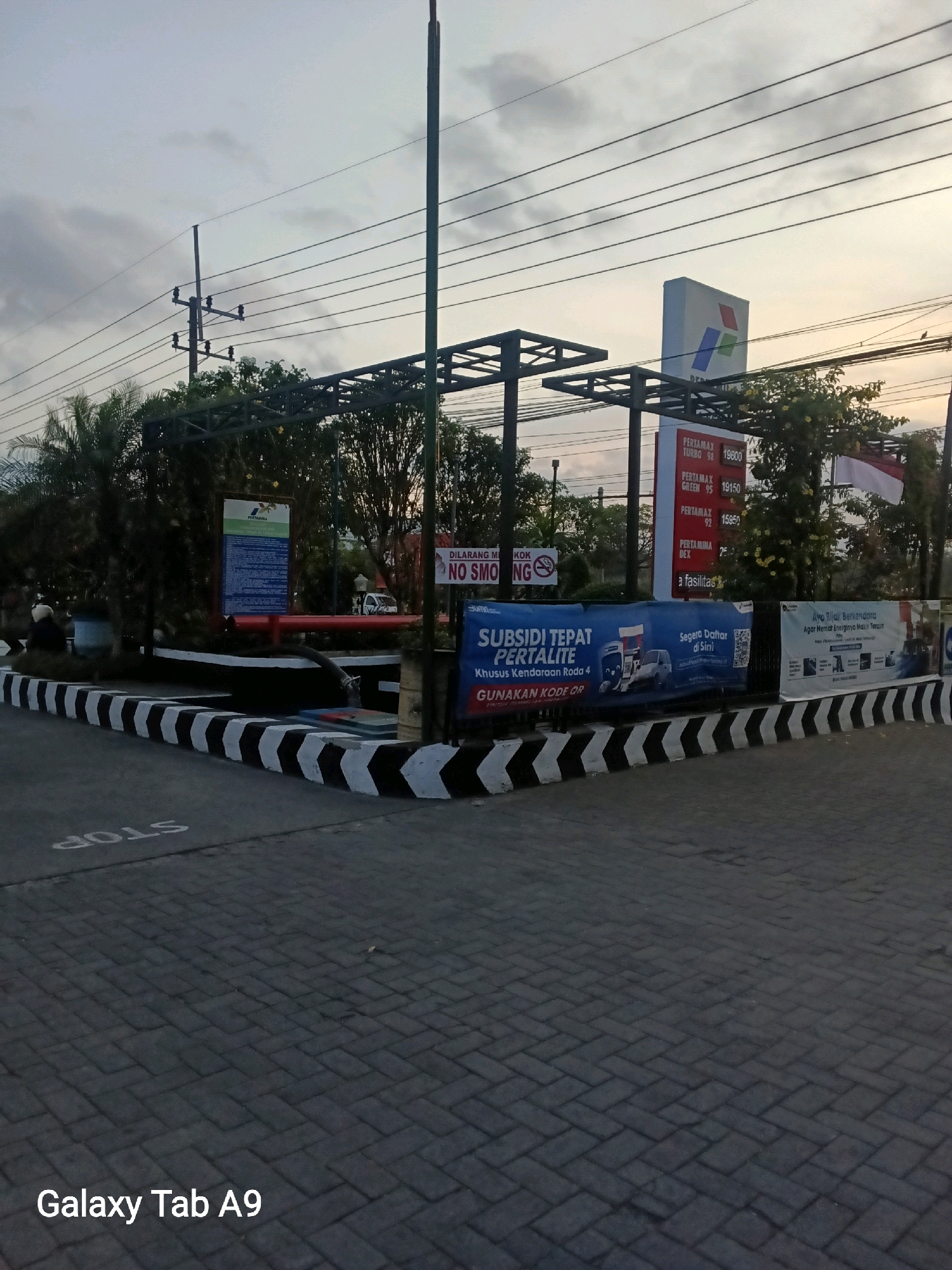The image depicts a Pertamina gas station in Trenggalek, Indonesia, at dusk. A large Pertamina sign stands prominently, displaying fuel prices. Billboards advertise "Subsidi Tepat Pertalite" and "Ayo Beli Kendaraan Agar Hemat Energi." The ground is paved with interlocking bricks, and a white "STOP" sign is painted on the road. Black and white chevron markings border the paved area. Electrical poles and overhead wires are visible. The sky is overcast, suggesting twilight. No people are clearly identifiable, though a figure sits on a bench near some palm trees.