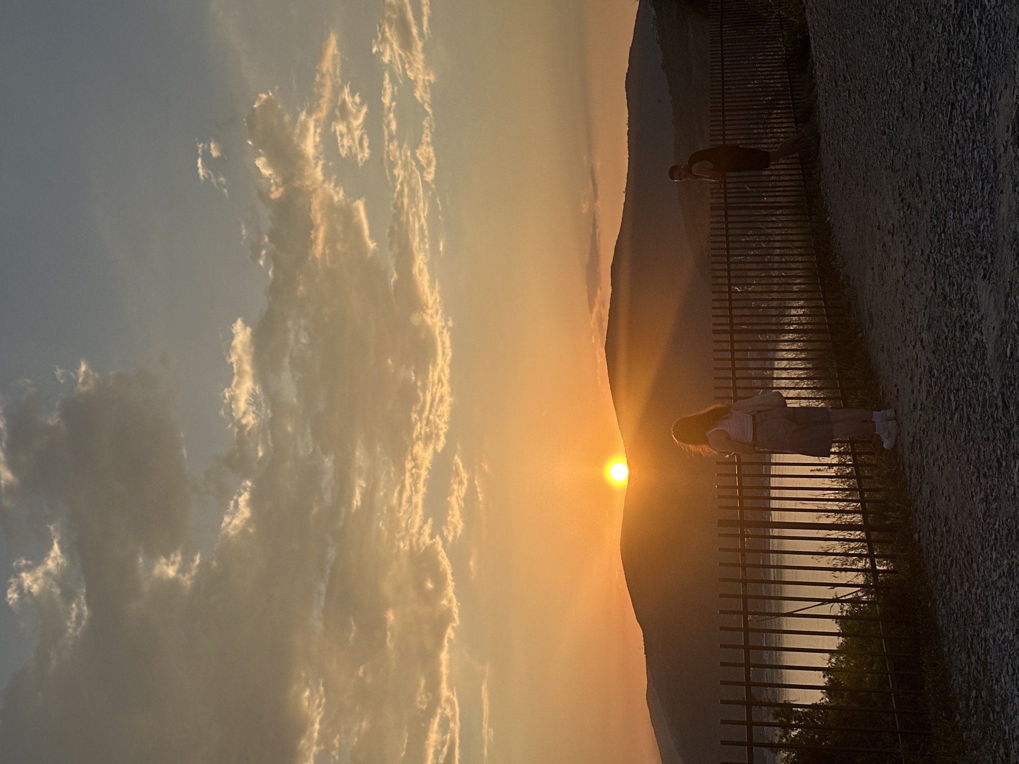 The image shows a sunset in Budva, Montenegro. The sun is setting behind a mountain, casting a golden glow over the sky and clouds. In the foreground, there is a fence and a path with two people standing nearby. One person is dressed in a light-colored shirt and dark shorts, while the other has long hair, a white shirt, and a denim dress.