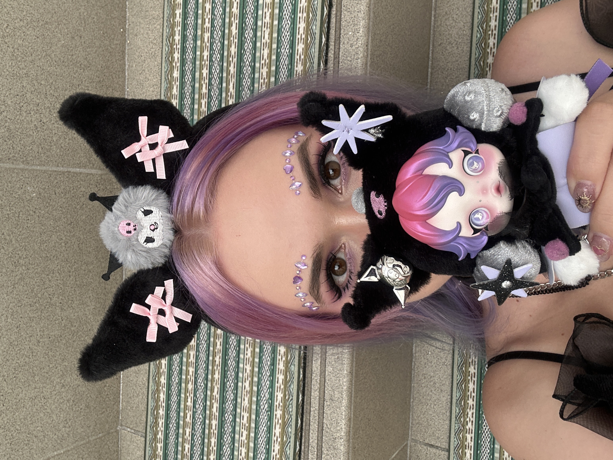A person with lavender hair is wearing a black headband with black, fuzzy bat ears adorned with pink bows and a grey puffball with a cartoon bat face. The person has facial gems applied in a line near their left eye. They are holding a black, fuzzy doll with a pink and purple hairstyle and large, wide-eyed face. The background features a vertically-oriented patterned rug or curtain and a stone or concrete wall, possibly indicating an interior setting, and steps leading up to a door. There is a gray stone background.