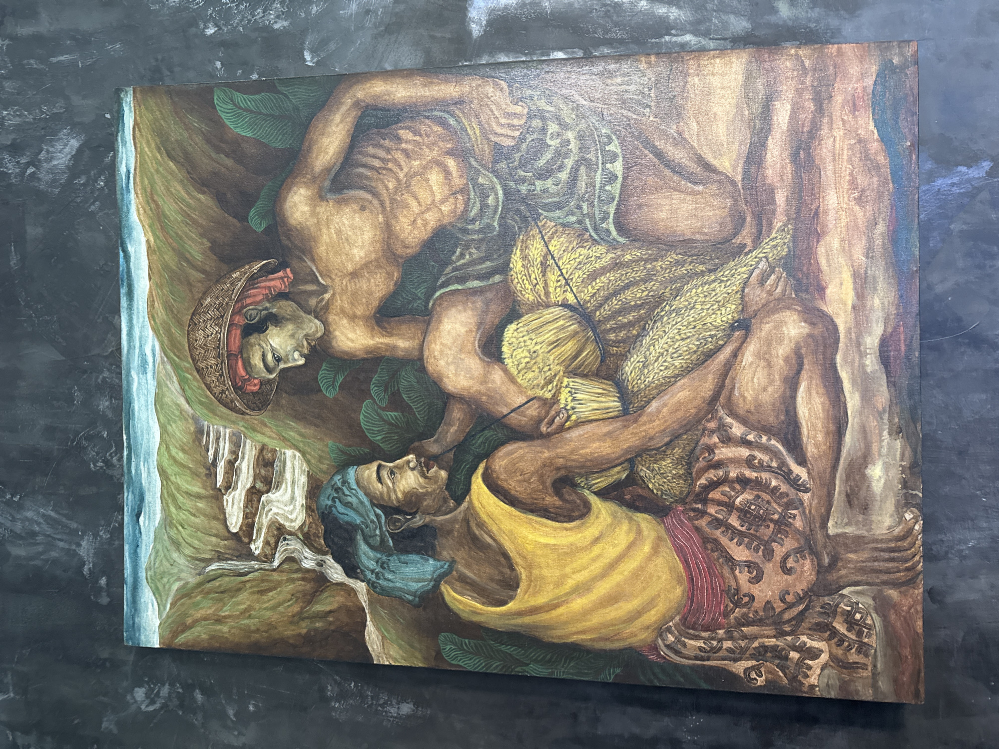 This is a vibrant, figurative painting depicting two male figures engaged in an agricultural activity, likely harvesting or carrying crops, set against a backdrop of natural elements. The art style features strong, somewhat stylized forms and a rich, earthy color palette.
The upper figure, positioned diagonally across the canvas, is a bare-chested man with a lean, muscular build. He wears a woven conical hat and patterned fabric around his waist. His gaze is directed downwards, focused on the task at hand. He appears to be holding or tending to large bundles of golden, harvested crops, possibly rice or wheat, which are tied with dark cords.
Below him, positioned along the bottom edge, is a second man. He wears a bright yellow sleeveless top and an intricately patterned sarong-like lower garment in shades of brown, red, and cream. A blue head covering adorns his head. He is depicted in a reclined or seated position, looking upwards with an open mouth, suggesting an expression of exertion, song, or perhaps shouting. His hands are also near or interacting with the bundles of crops.
The background on the left features a strip of light blue water, indicative of an ocean or river, bordered by a green-brown cliff face with white, waterfall-like formations. Lush green foliage and tree elements are scattered throughout the composition, reinforcing a tropical setting. The right side of the painting shows more abstract tree trunks or rock formations in brown and dark tones.
The painting conveys a sense of laborious work and connection to nature, characteristic of agricultural life. There is no visible text in the image. Given the context, this scene likely depicts a moment from daily life or traditional harvest in a place like Sukawati, Indonesia.